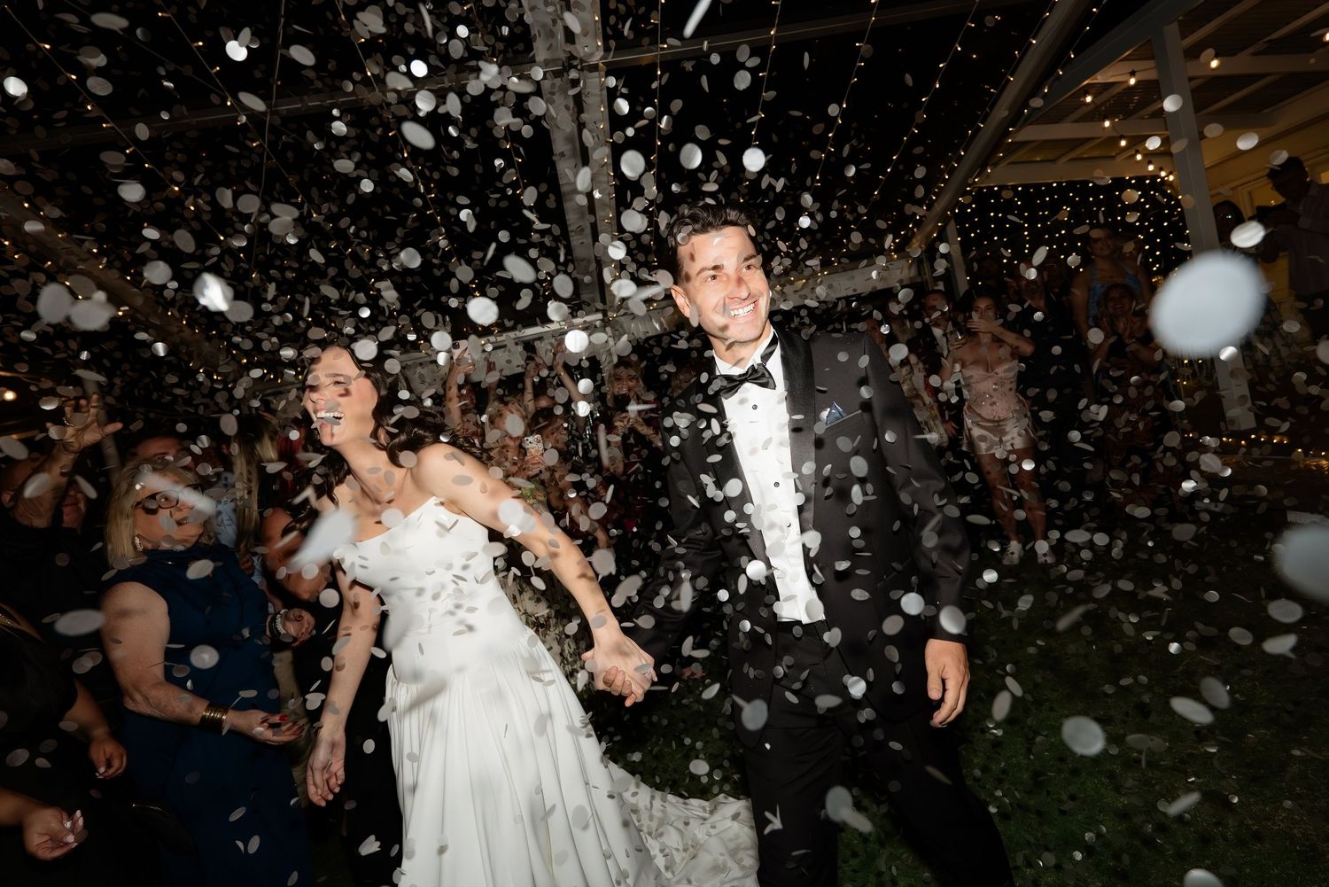 Bride and groom celebrate joyfully as confetti showers over them during their festive outdoor wedding exit.