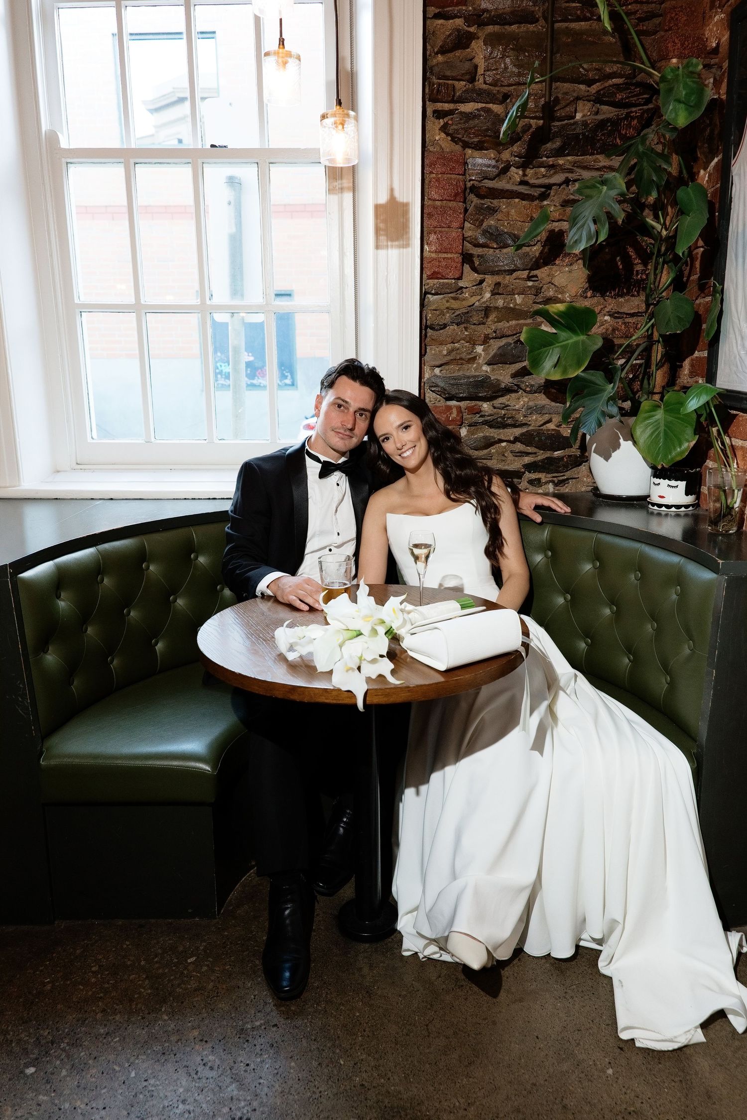 Bride and groom sitting together on a green tufted booth by a window in a rustic venue.