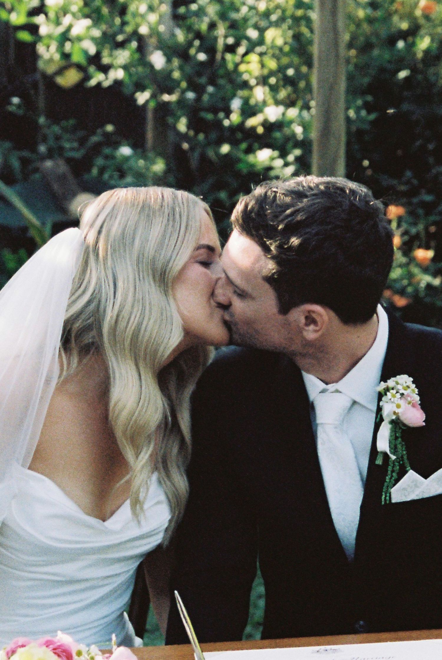 Bride and groom sharing a kiss at the signing table surrounded by lush greenery on their wedding day.