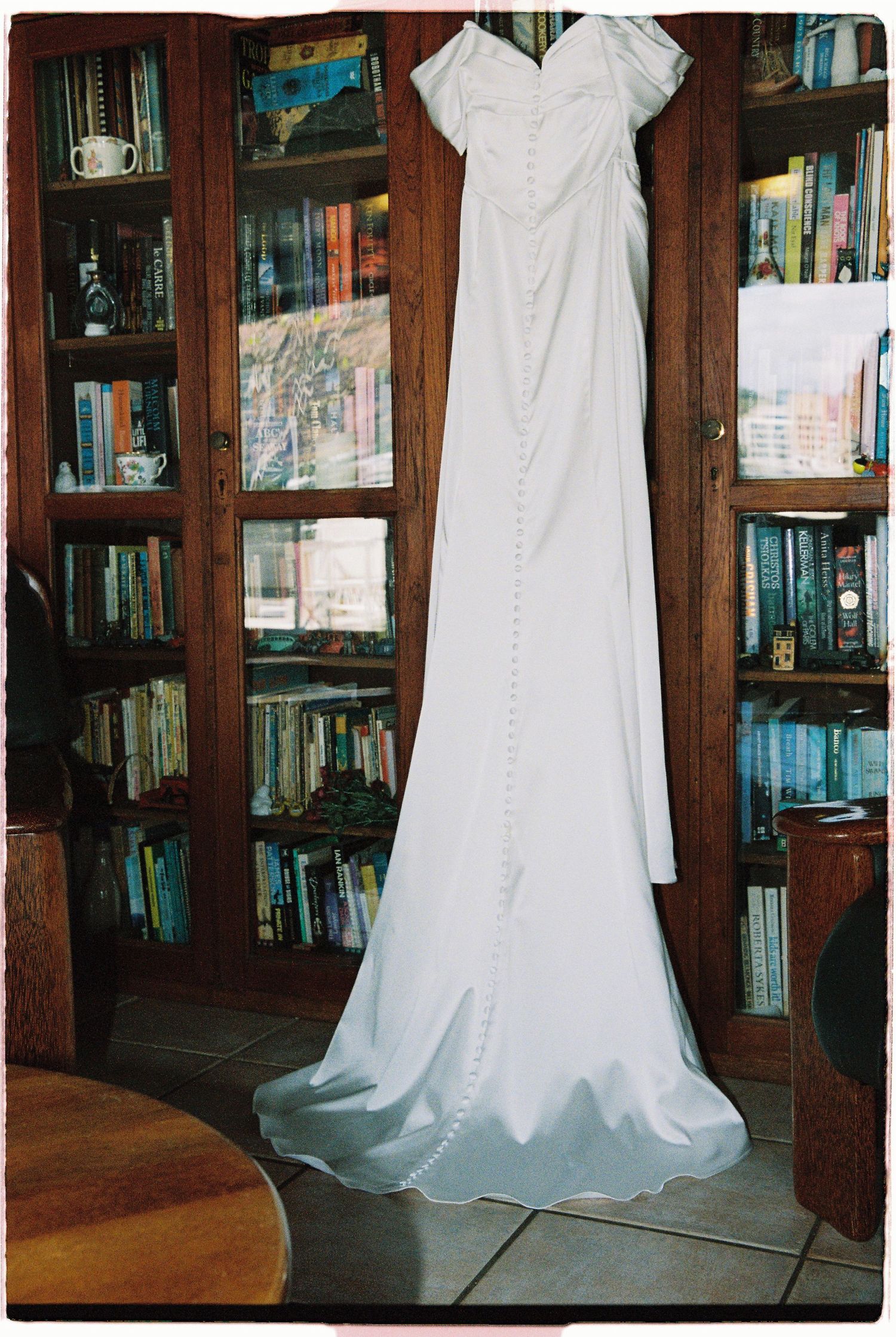 Elegant white off-shoulder wedding gown hanging in front of wooden bookshelves filled with books.