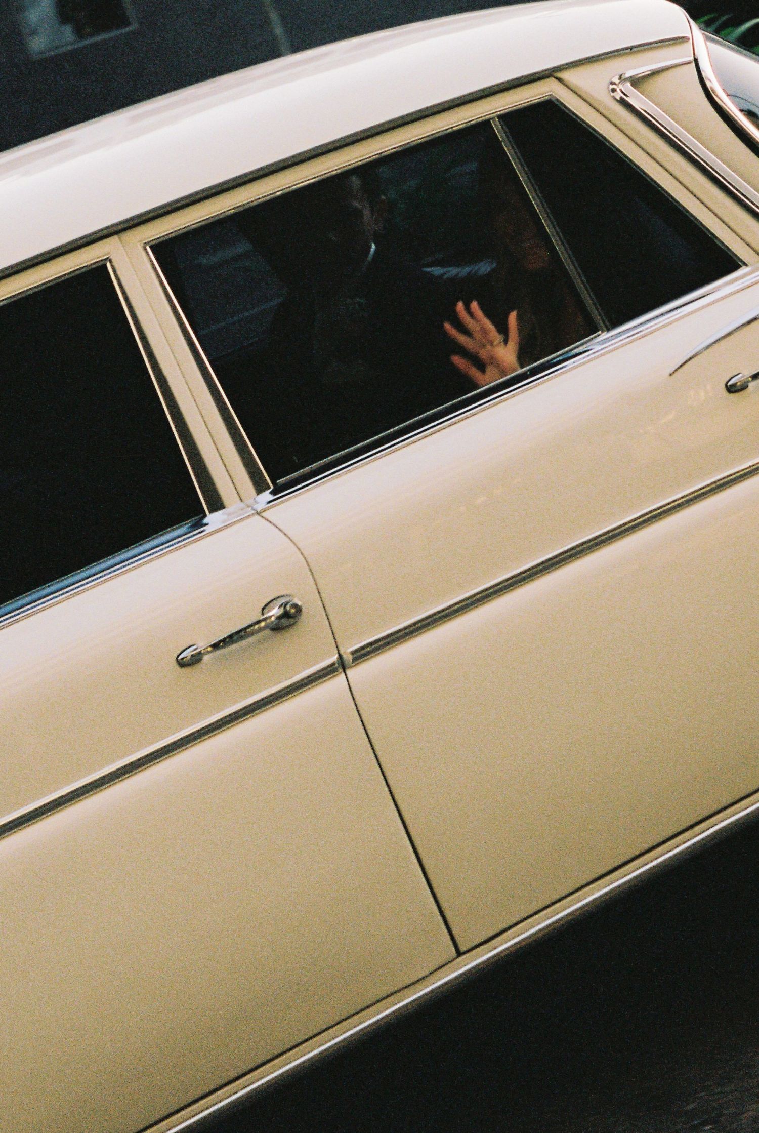 Bride waving from window of vintage cream-colored classic car on wedding day.