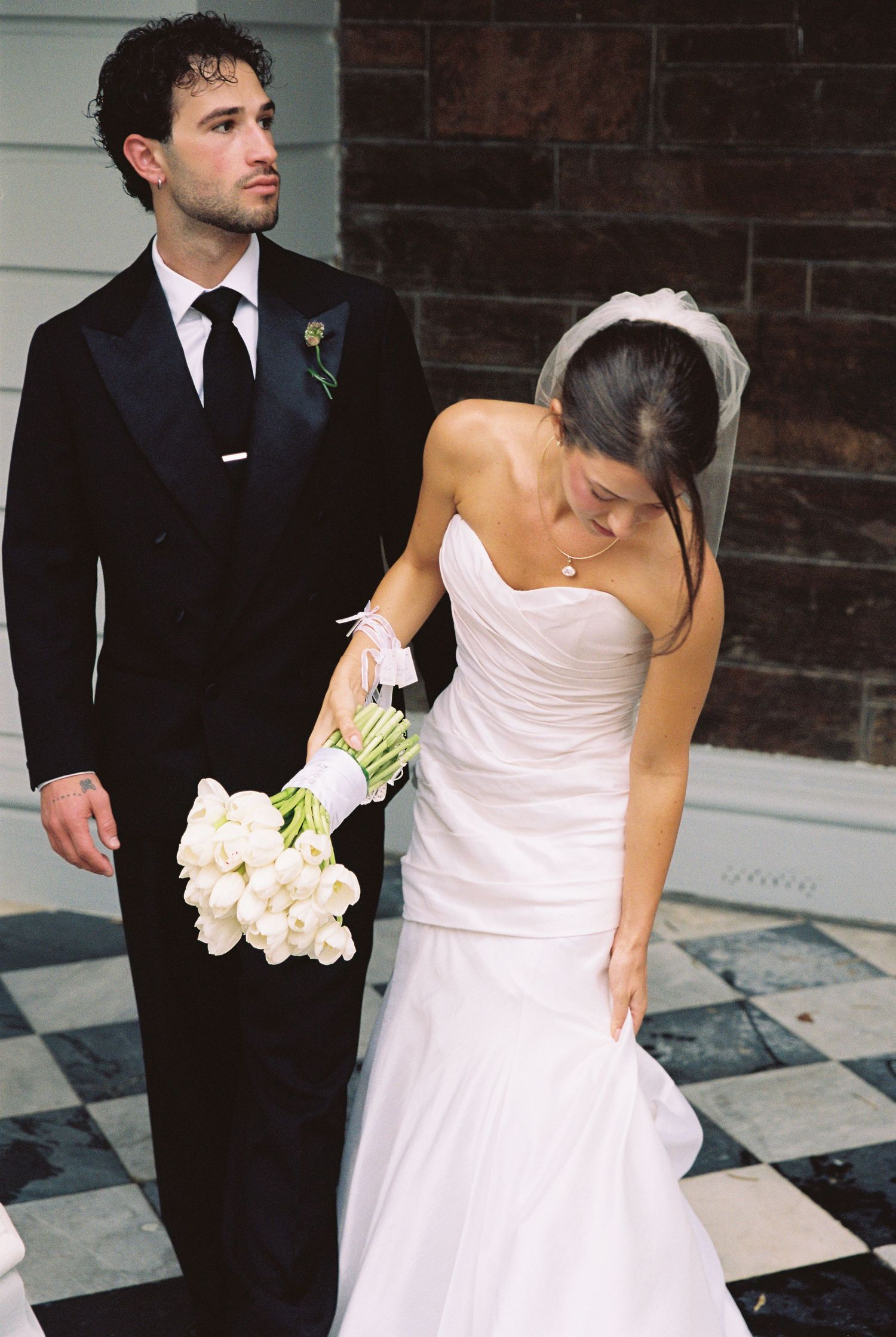 Groom in black tuxedo holds white tulip bouquet as bride in strapless gown adjusts her dress.