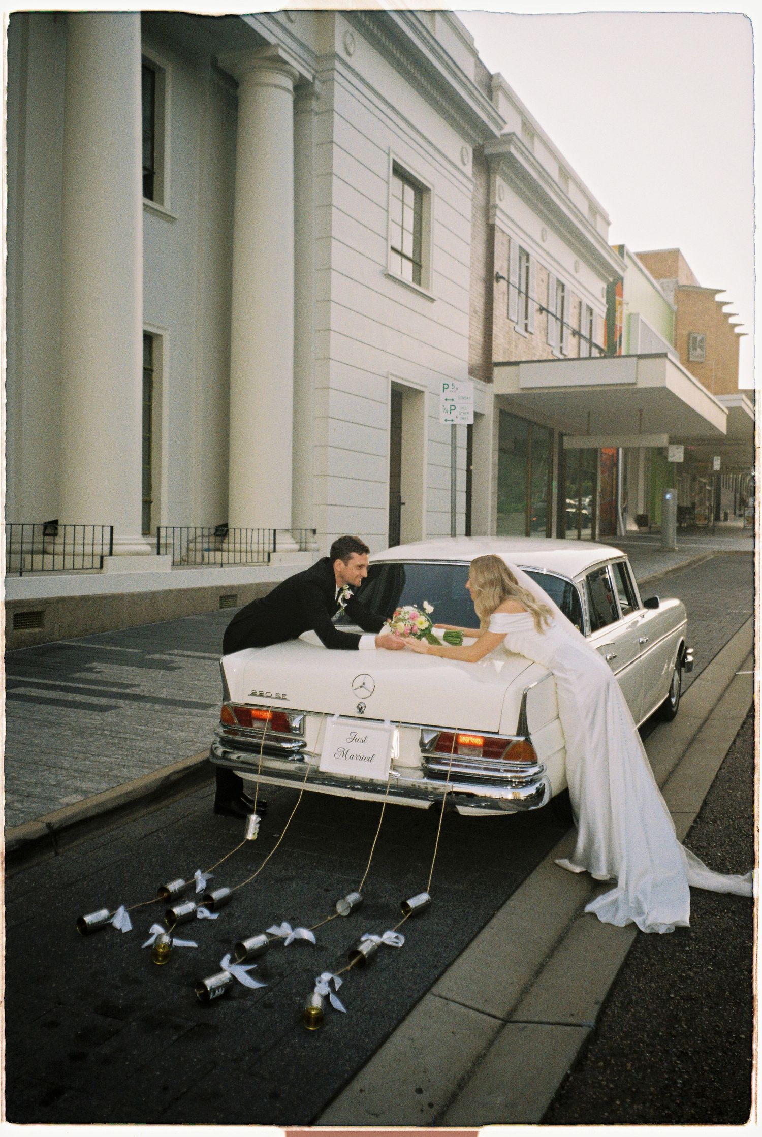 Bride and groom leaning over the trunk of a vintage white Mercedes wedding car with tin cans trailing.