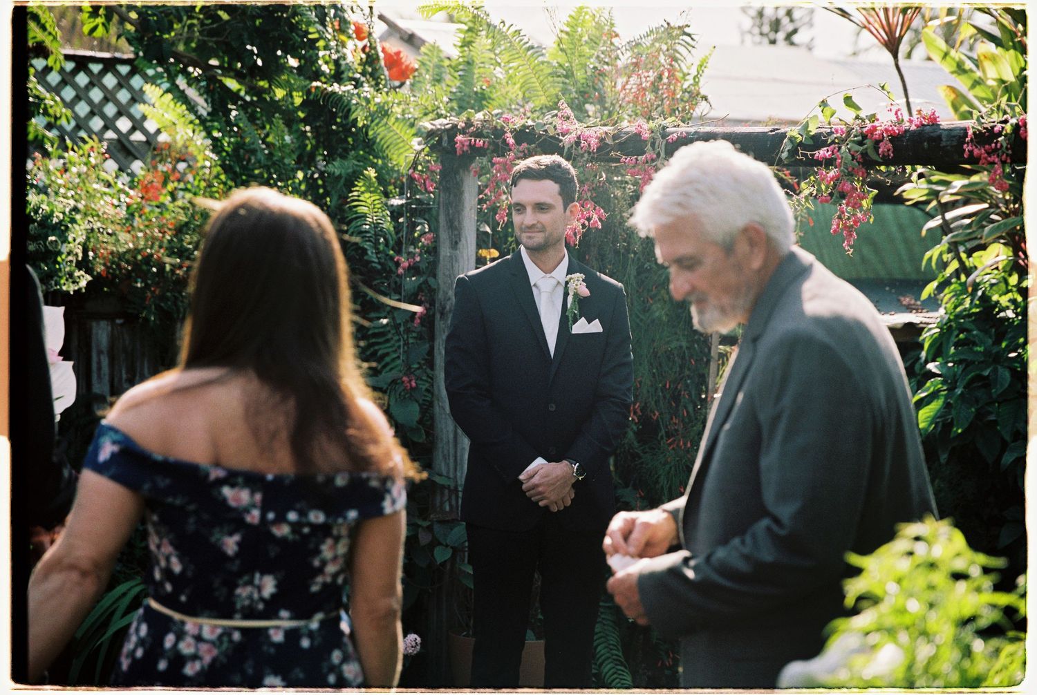 Groom in black tuxedo stands in lush garden ceremony space as guests gather for outdoor wedding.
