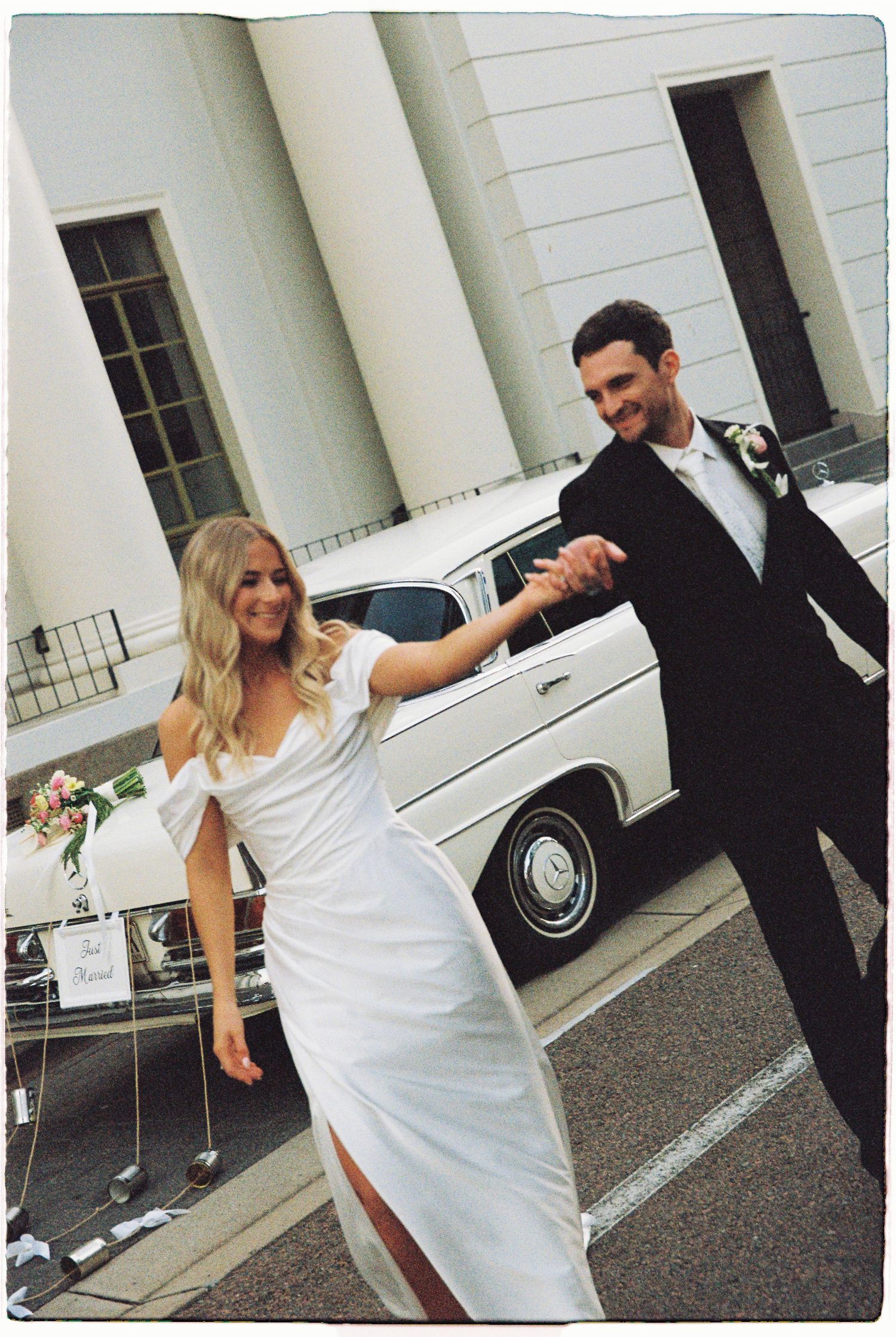 Newlywed couple holding hands and dancing joyfully outside wedding venue beside vintage white car.