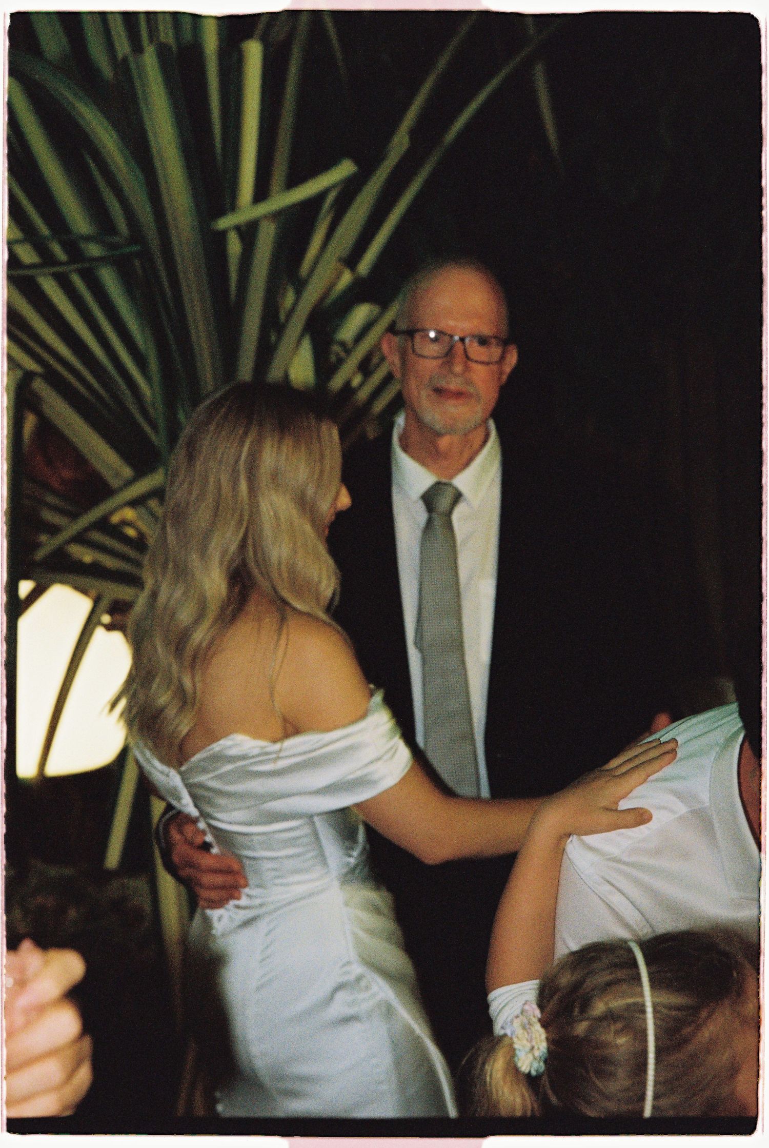 A bride in a white off-shoulder gown embraces an older man in glasses at an evening wedding celebration.