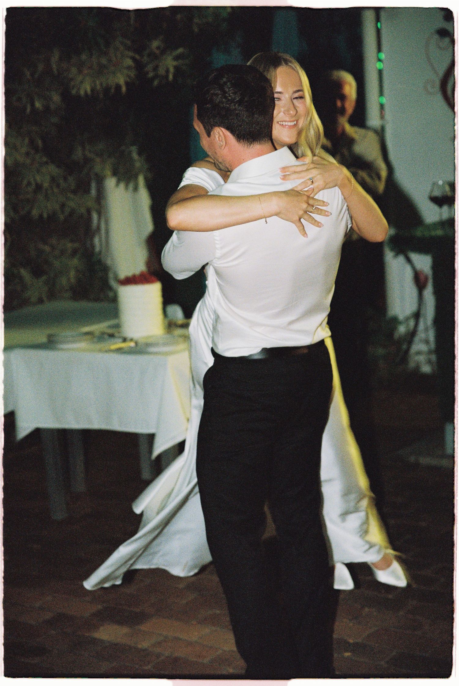 Bride and groom sharing first dance embrace at wedding reception with guests watching in background.