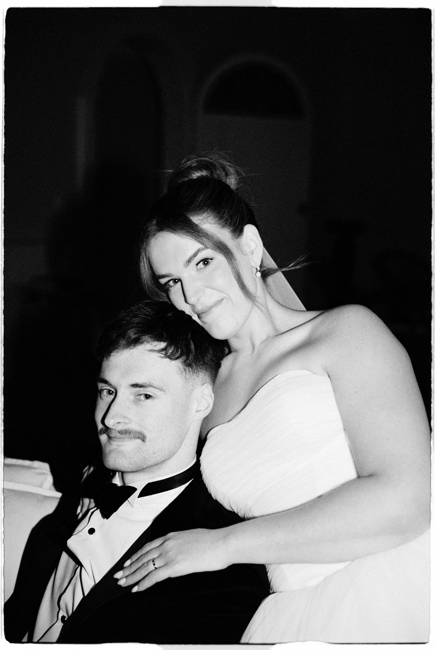 Black and white wedding portrait of bride leaning over seated groom in tuxedo indoors.