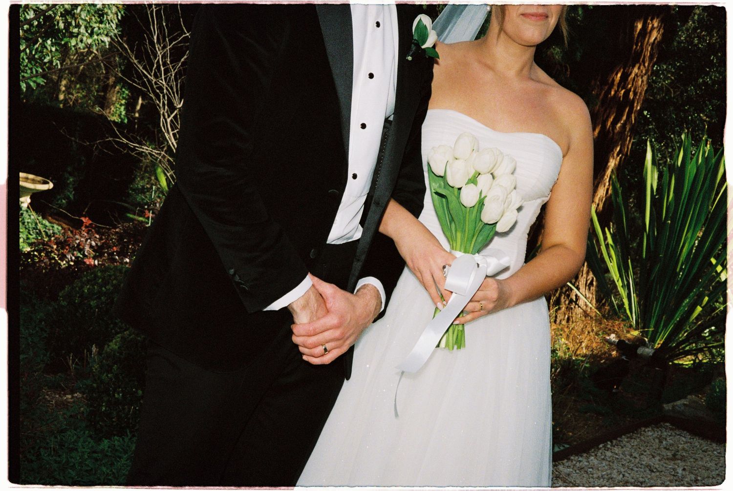 Bride holds white tulip bouquet while groom holds her hand in garden.