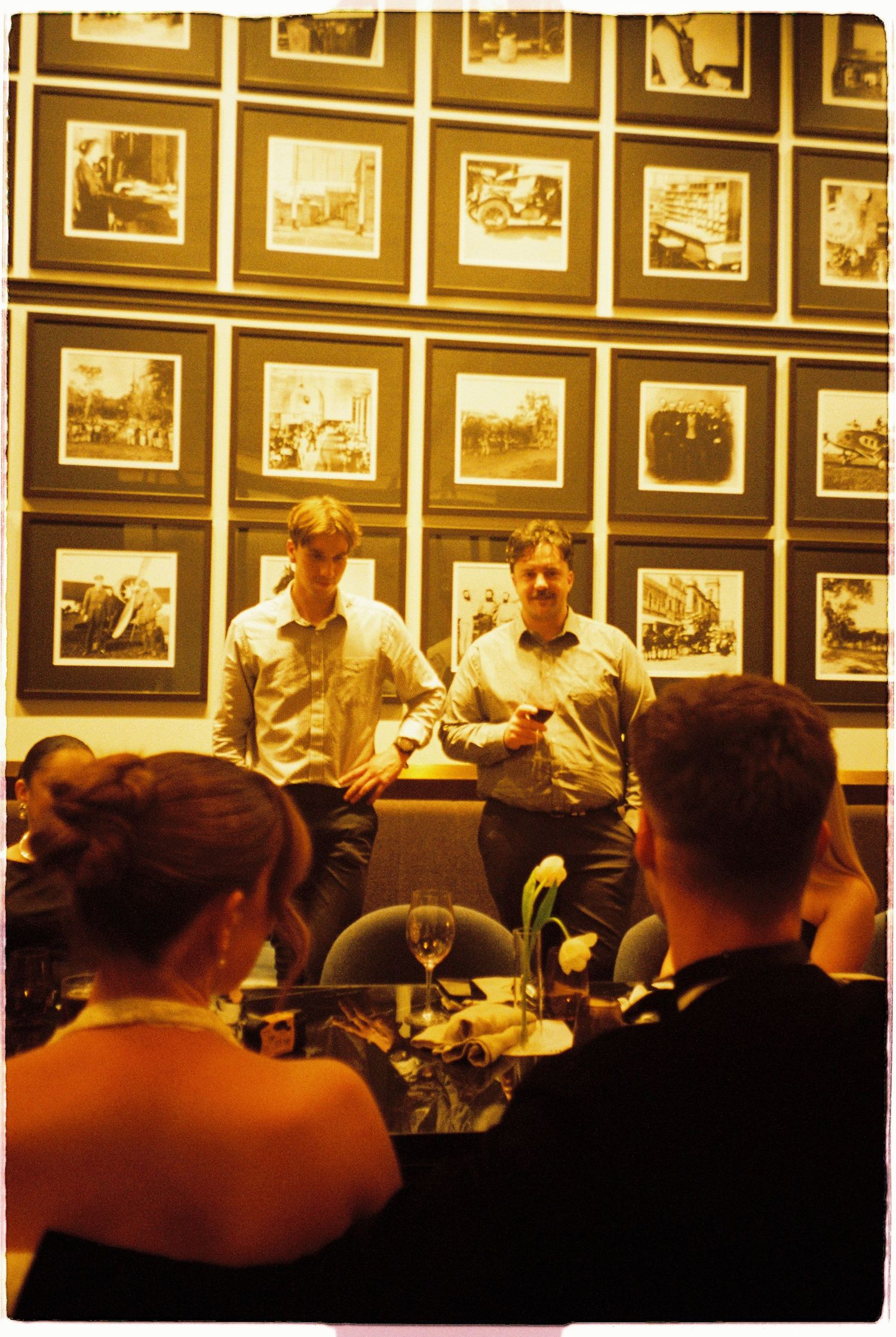 Two men in white shirts speak to seated wedding guests in dimly lit restaurant with framed photos on wall.