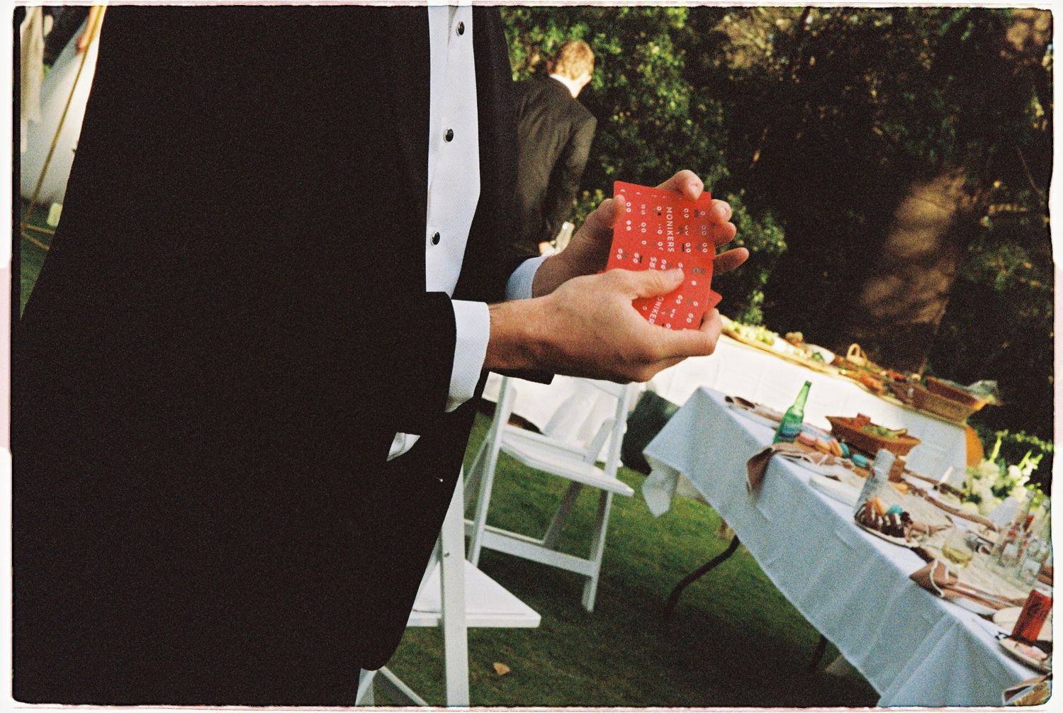 Groom in tuxedo holds a red gift at an outdoor wedding reception with white chairs.