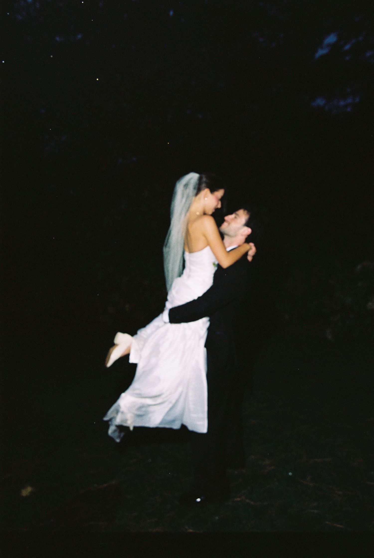 Groom lifting bride in white strapless gown and veil in romantic nighttime wedding celebration portrait.