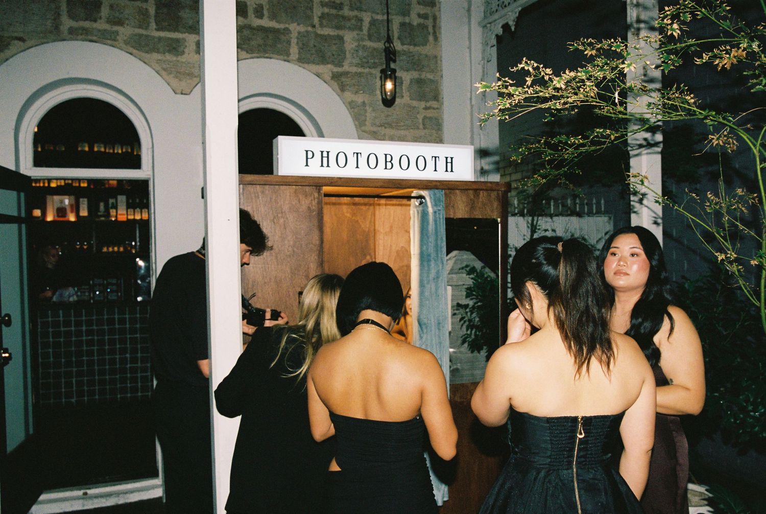 Wedding guests in formal attire queuing outside a photo booth at an evening reception.