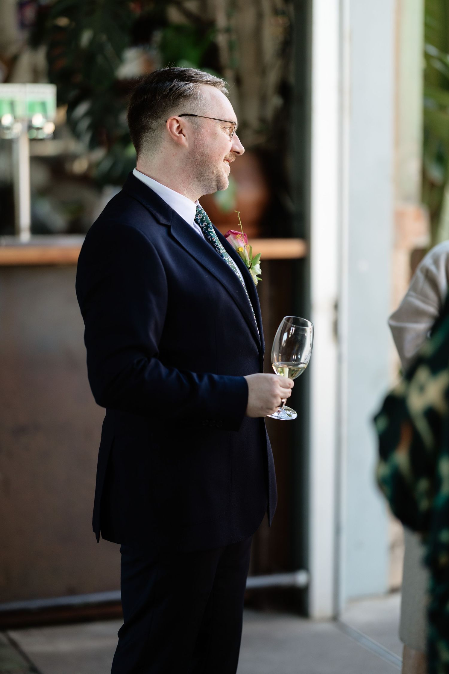 A well-dressed man in a dark suit with boutonniere holds a wine glass at an elegant wedding reception.