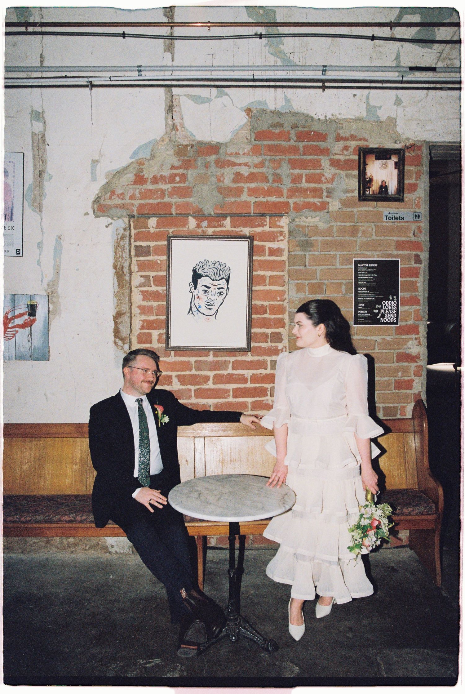 A bride in white dress and groom in dark suit sit at a small cafe table against a rustic brick wall.