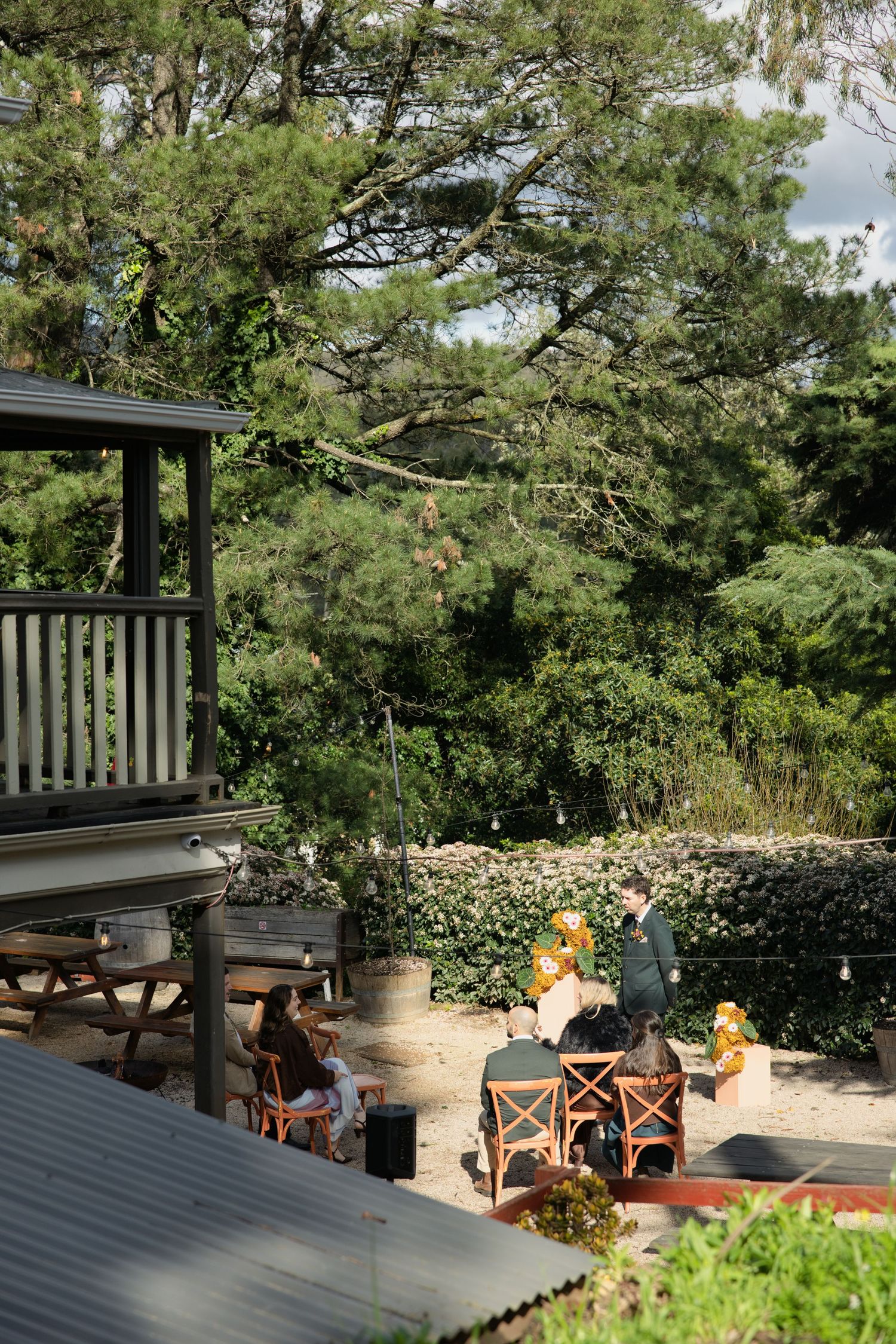 People dining outdoors at wooden tables in a lush garden setting with tall trees and green hedges.