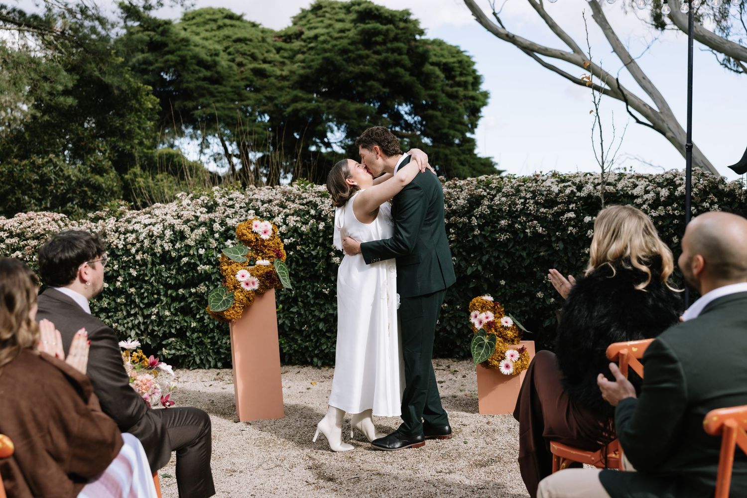 Bride and groom share first kiss at outdoor wedding ceremony with floral arrangements and seated guests.