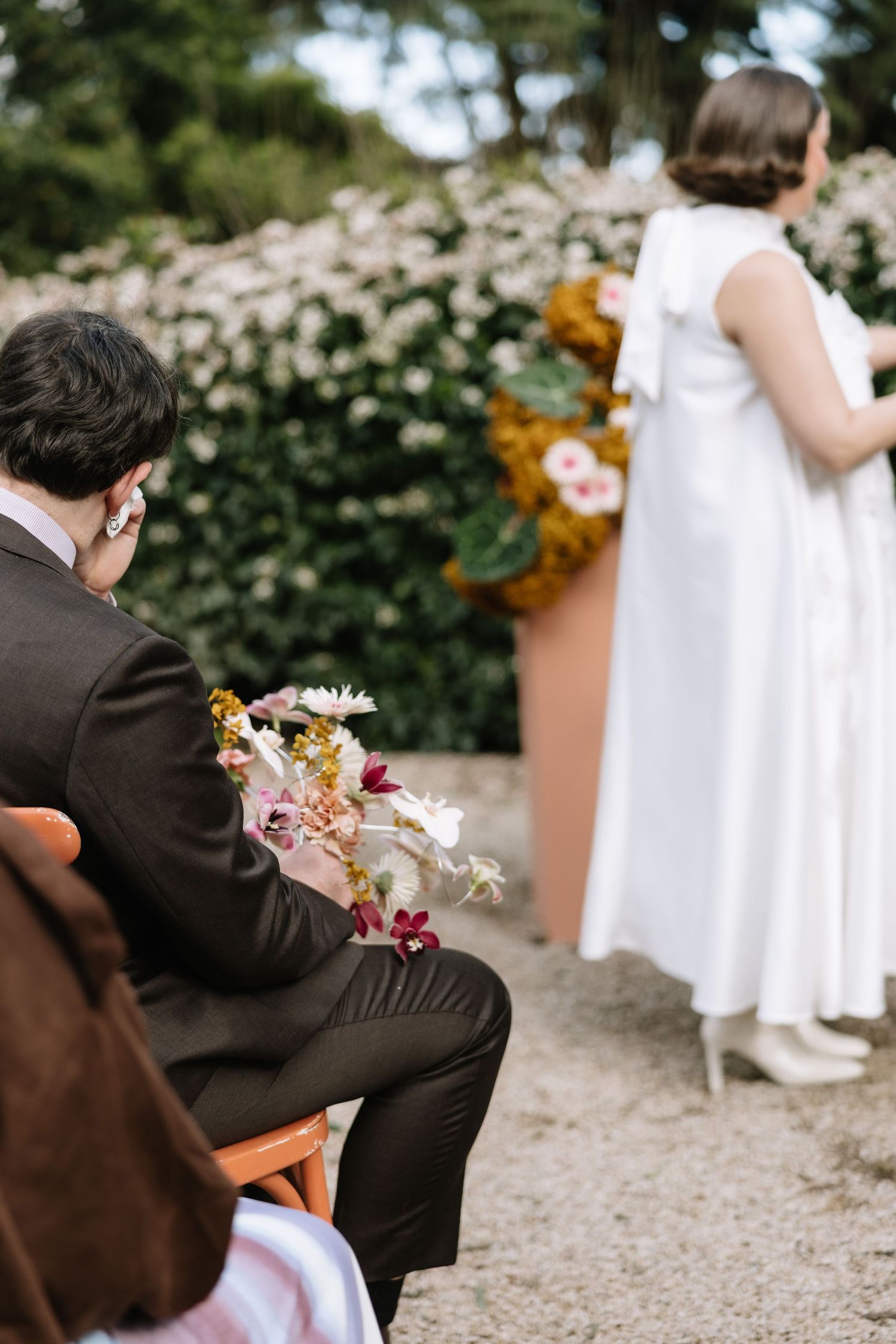 A groom in a brown suit holds a floral bouquet while seated at an outdoor wedding ceremony with a bride in white ahead.