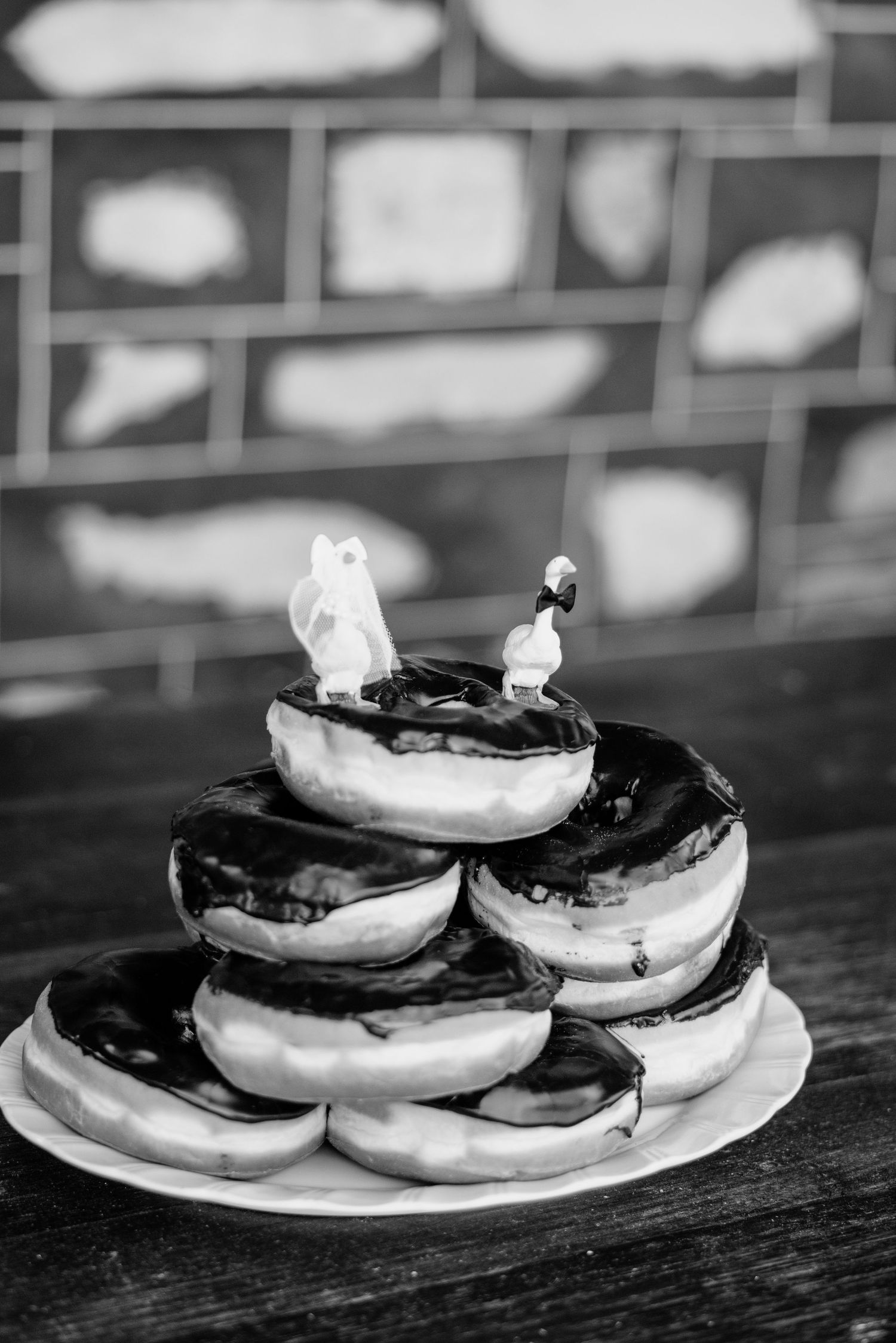 Black and white photo of stacked chocolate glazed donuts topped with miniature bride and groom wedding cake figurines.