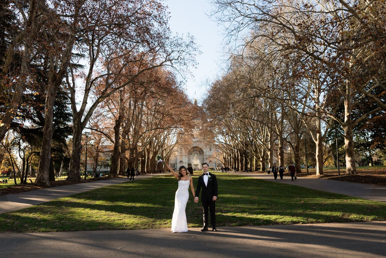 Couple in wedding attire standing on a tree-lined path with a historic building backdrop in a lush green park.