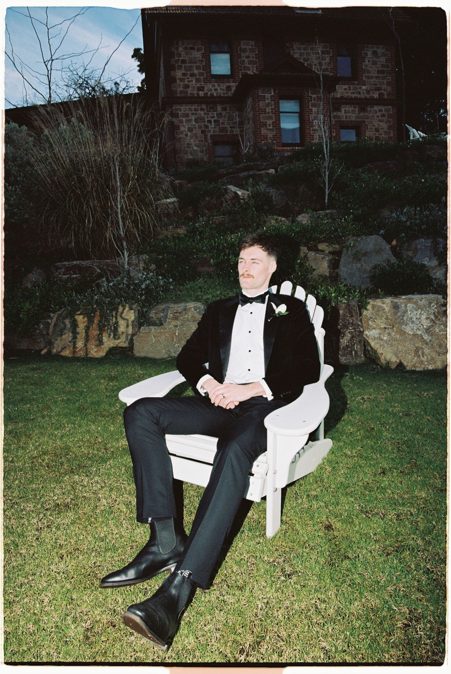 Groom in black tuxedo relaxing on white Adirondack chair outdoors with stone building behind him.