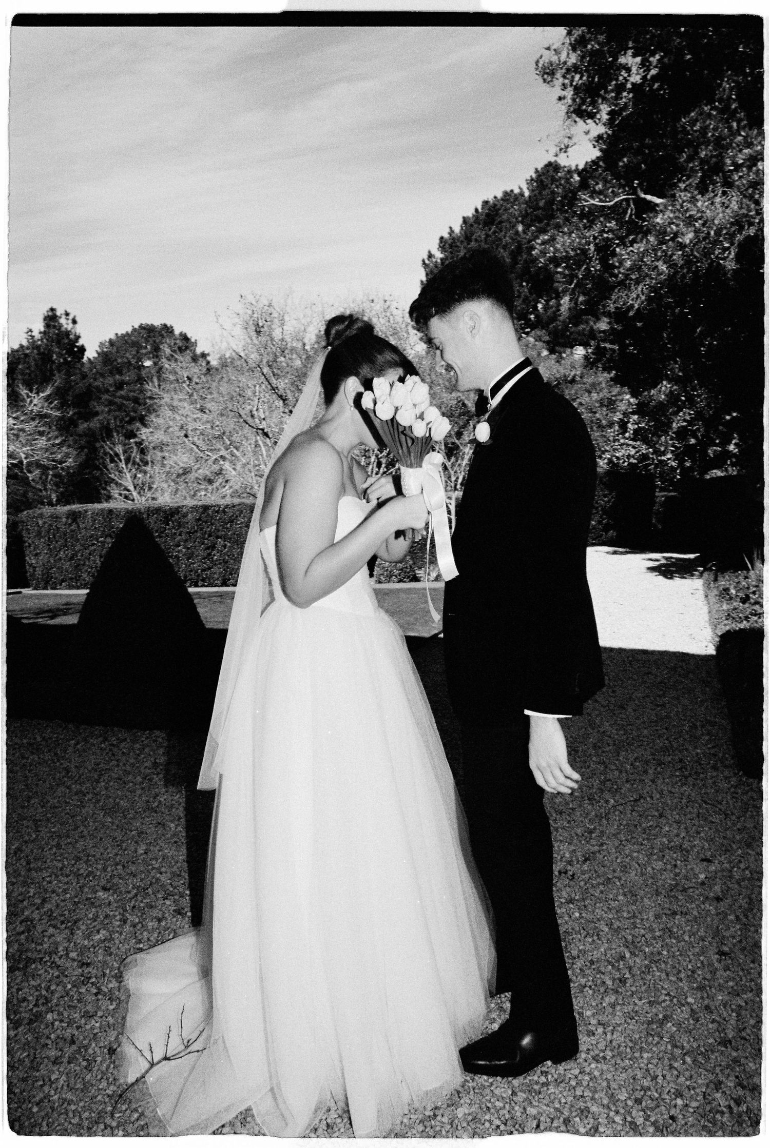 Black and white photo of a bride and groom sharing a romantic moment with a floral bouquet at an outdoor wedding.