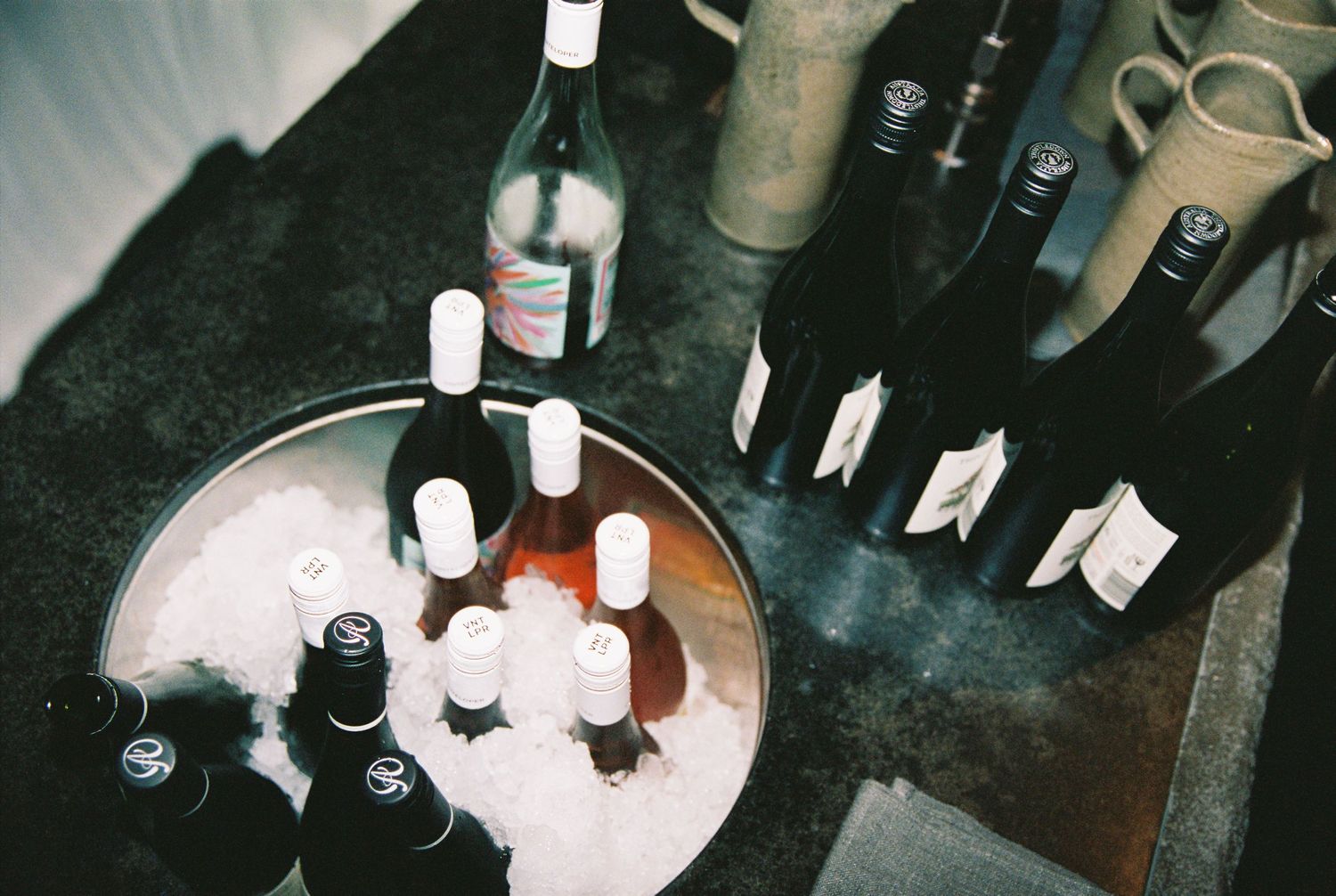 Assorted wine bottles chilling in an ice bucket alongside additional bottles on a dark surface at a wine tasting event.