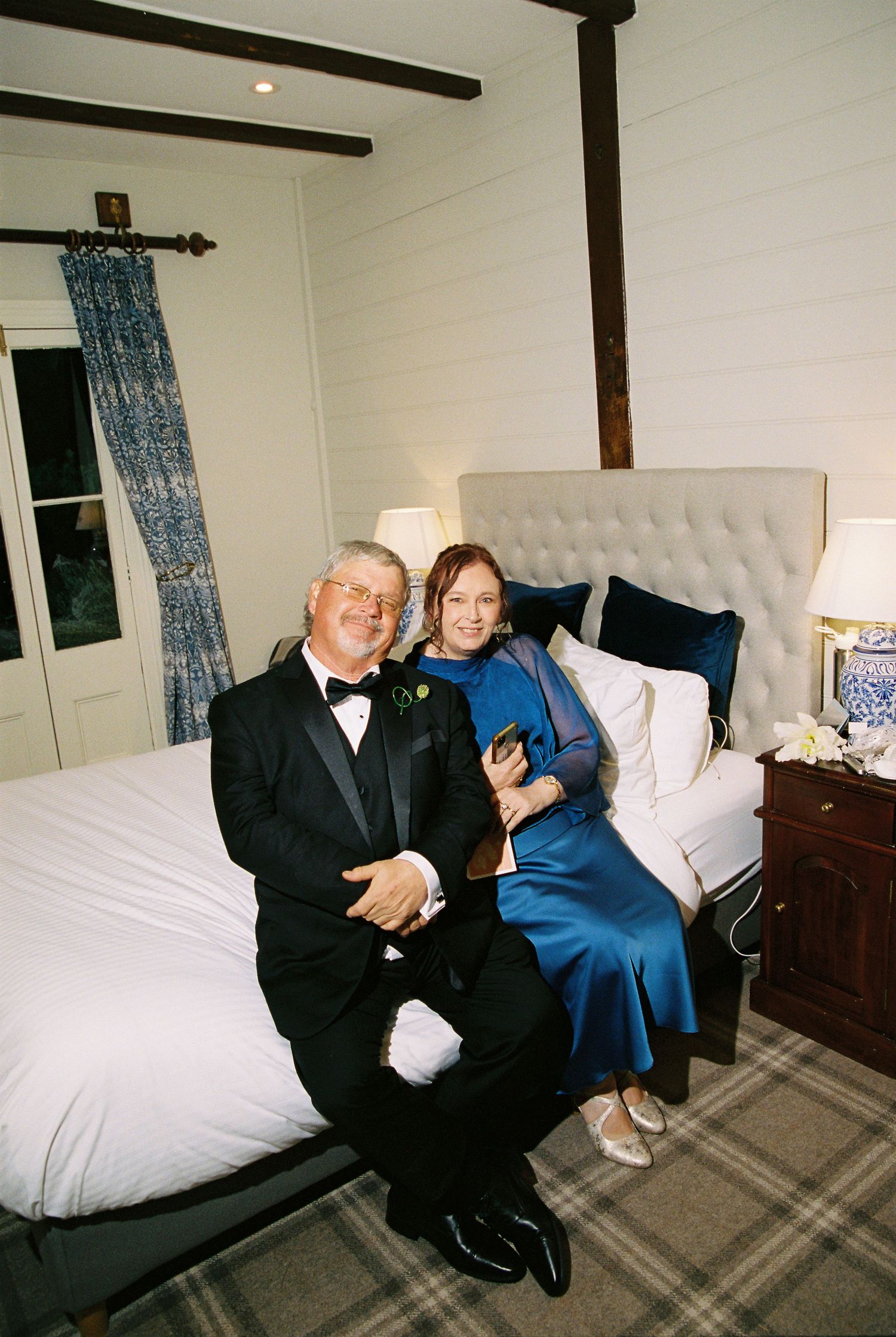 Couple in formal wedding attire sits on tufted bed in elegant bedroom, smiling warmly together.