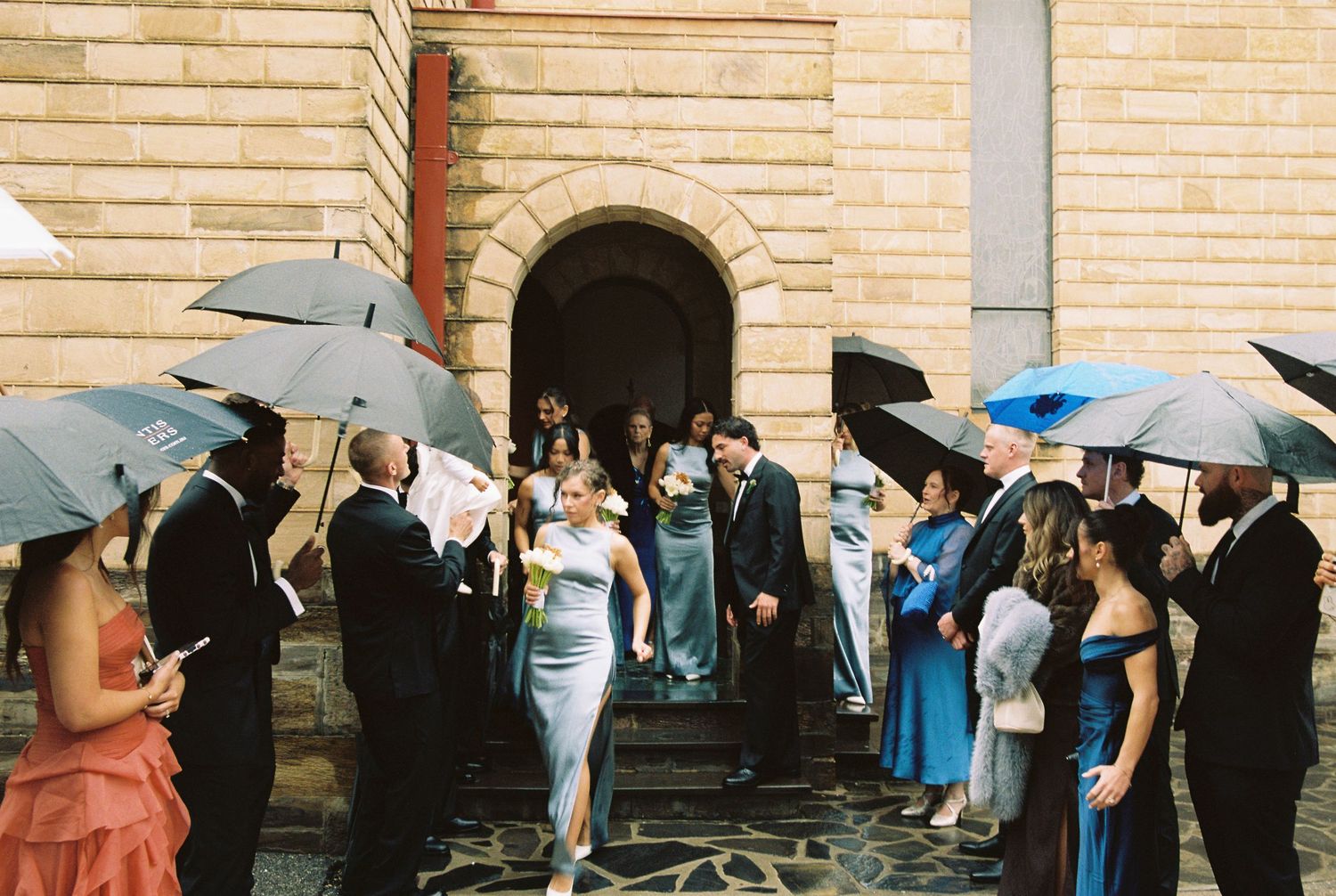 Wedding guests holding umbrellas outside a stone church entrance as bride and groom descend the steps.