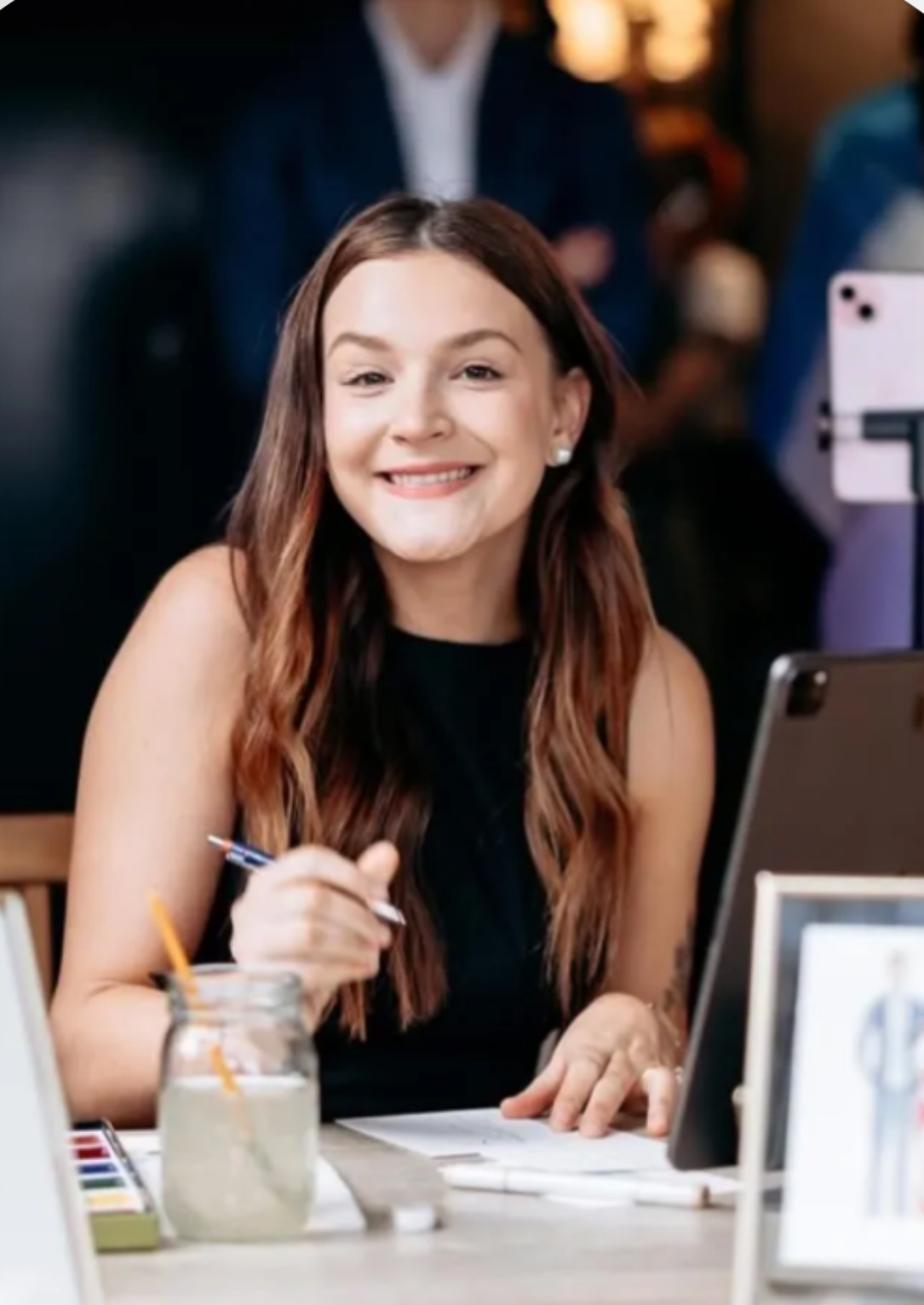 Smiling young woman with long brown hair sitting at a desk with watercolors and a tablet, holding a pen.