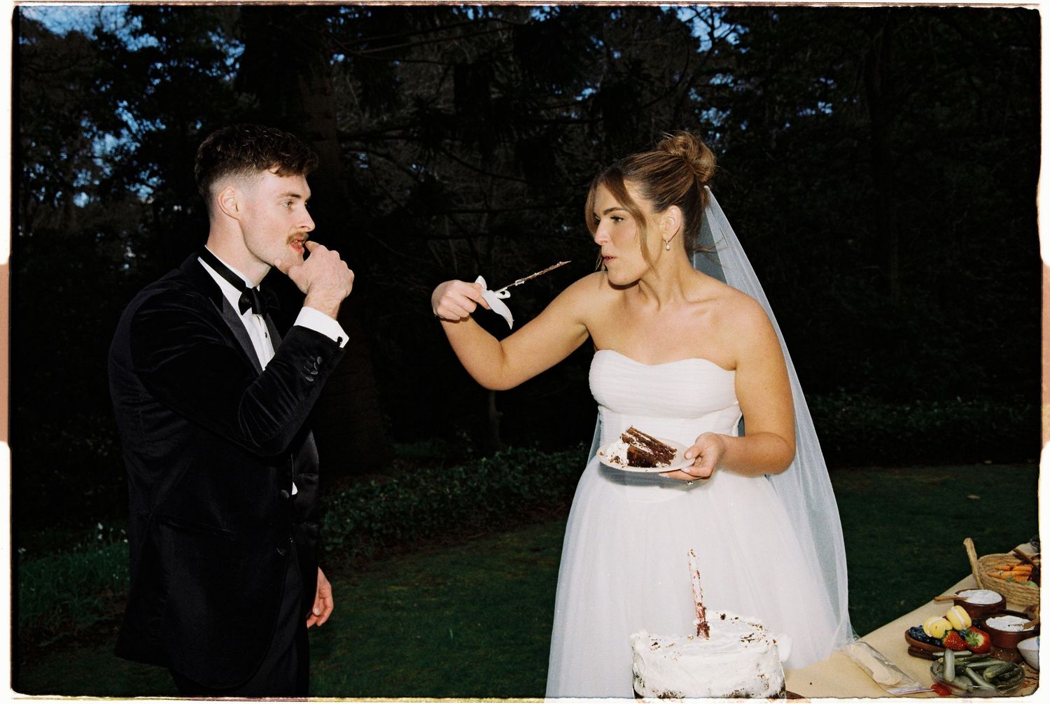 Bride feeding wedding cake to groom in black tuxedo at outdoor evening wedding reception.