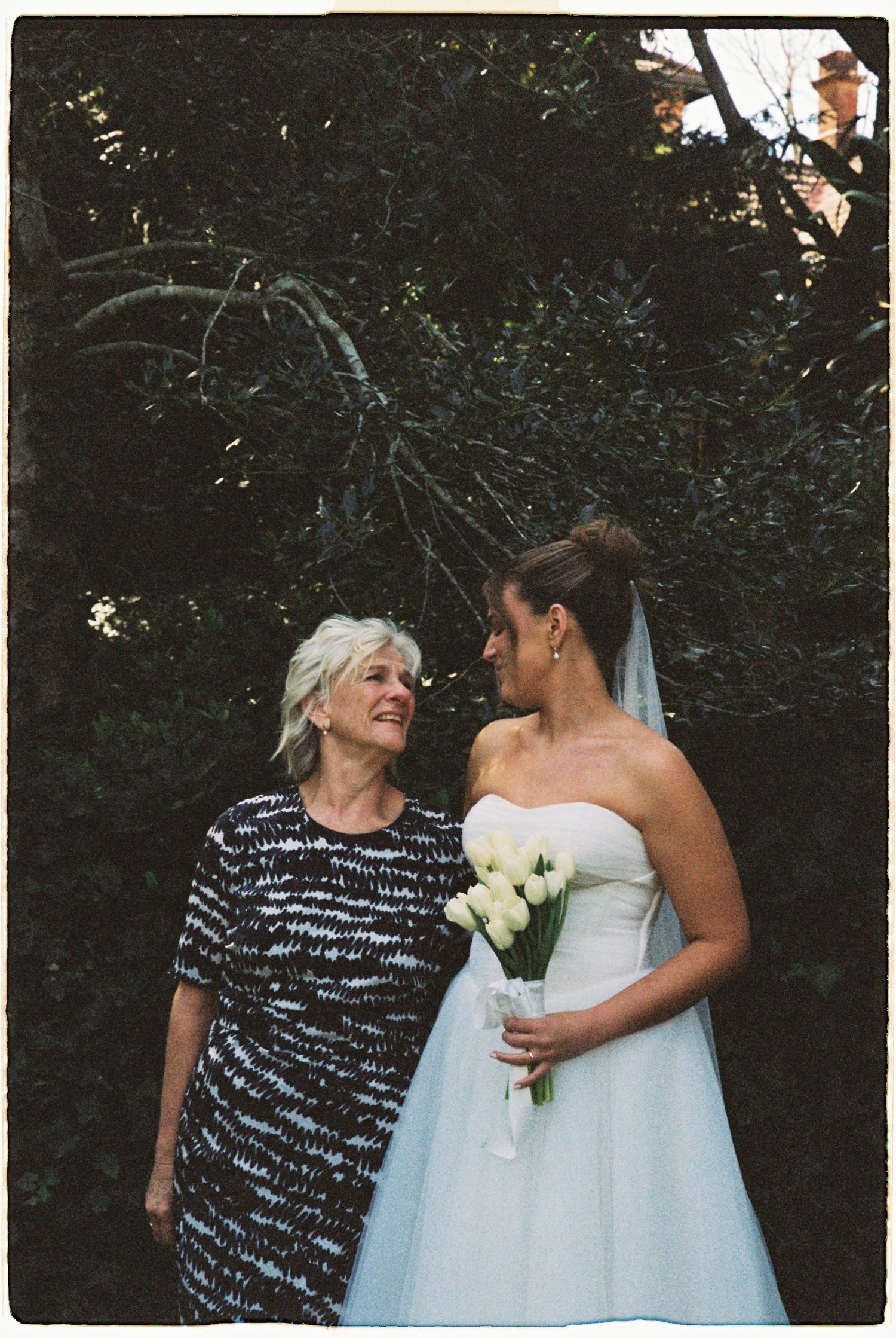 Bride in strapless white gown holding white tulip bouquet shares tender moment with older woman.