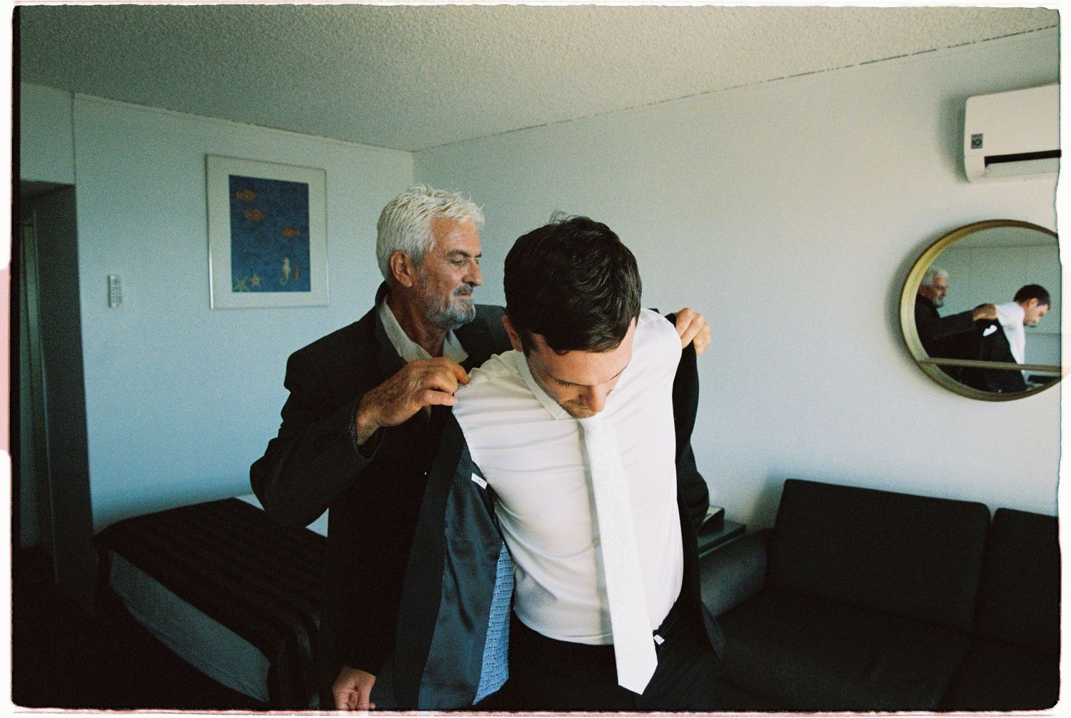 Older man helping a groom put on his suit jacket in a hotel room before a wedding.