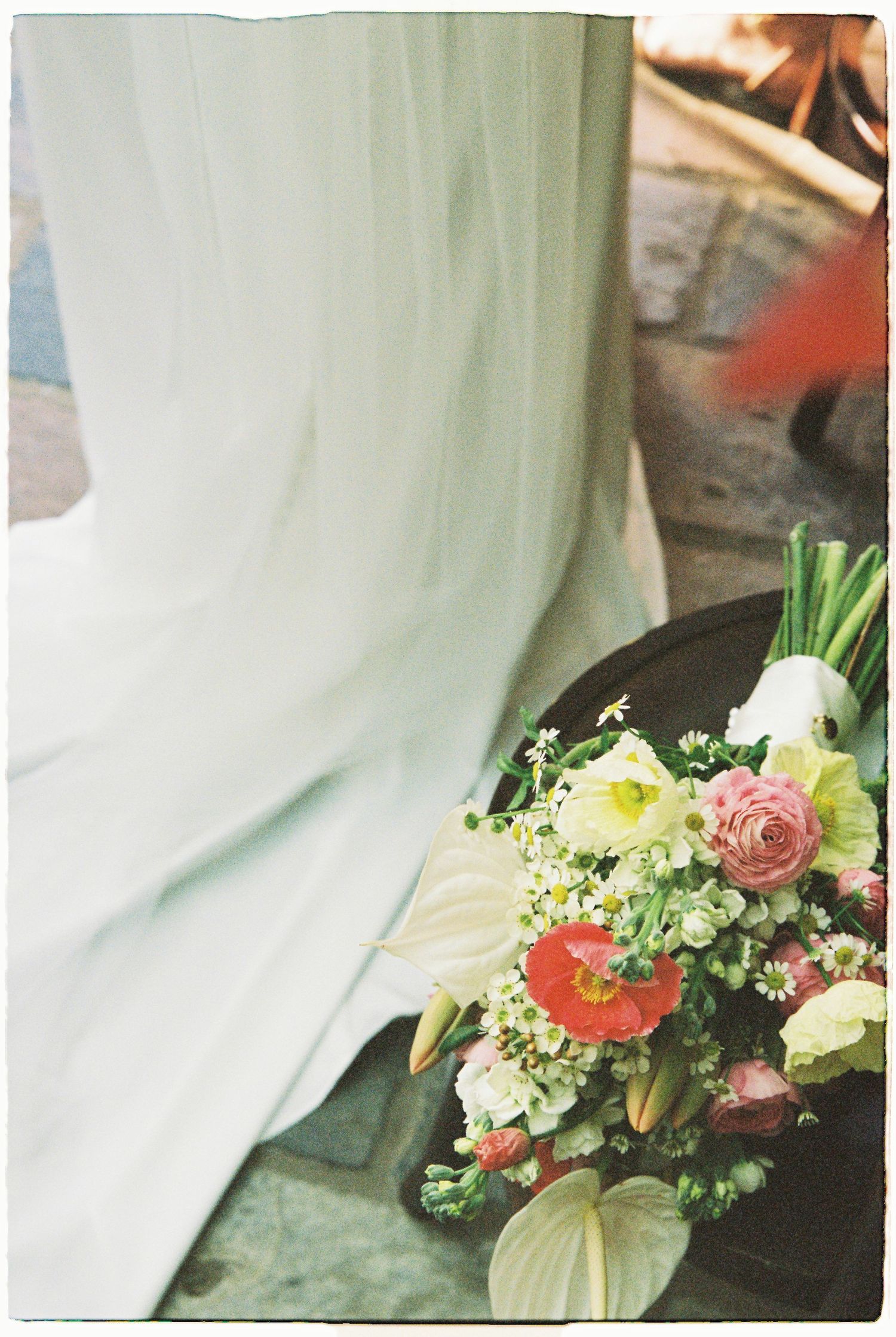 Colorful bridal bouquet with pink roses, red anthuriums, and white flowers resting beside a white wedding gown.