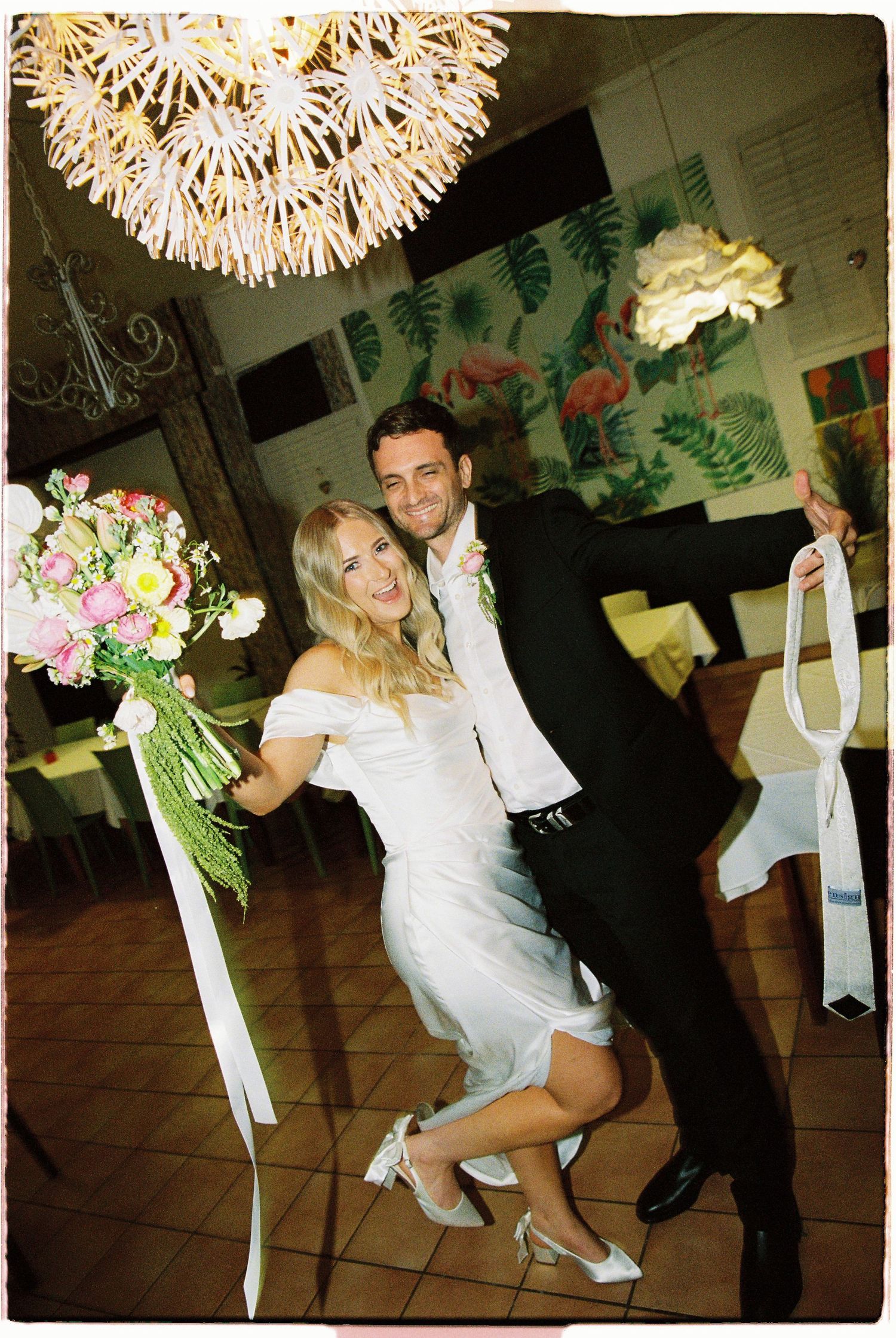 Bride and groom pose playfully indoors under decorative chandelier with wedding bouquet.