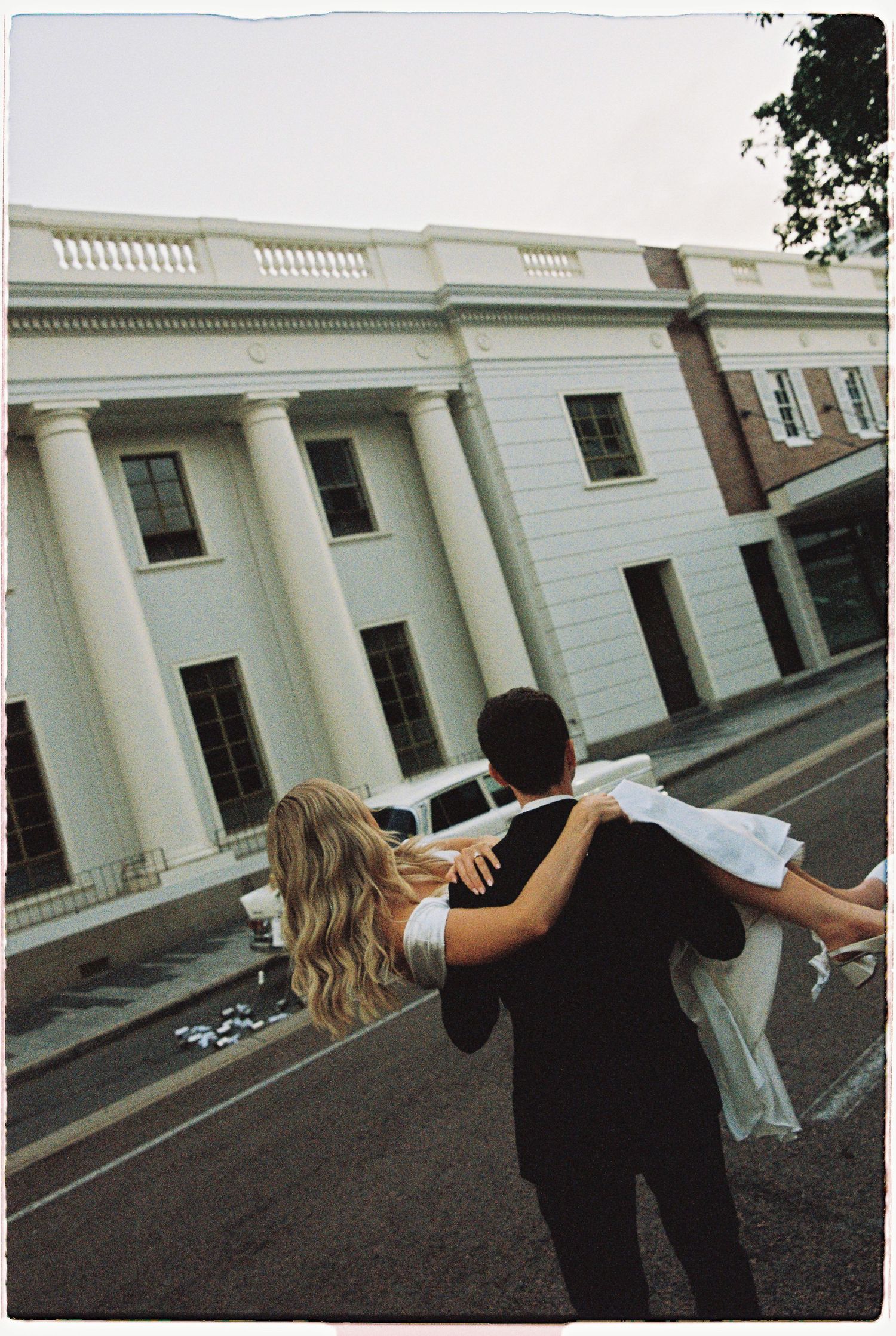 Groom carries bride in wedding dress toward a grand white columned building on their wedding day.