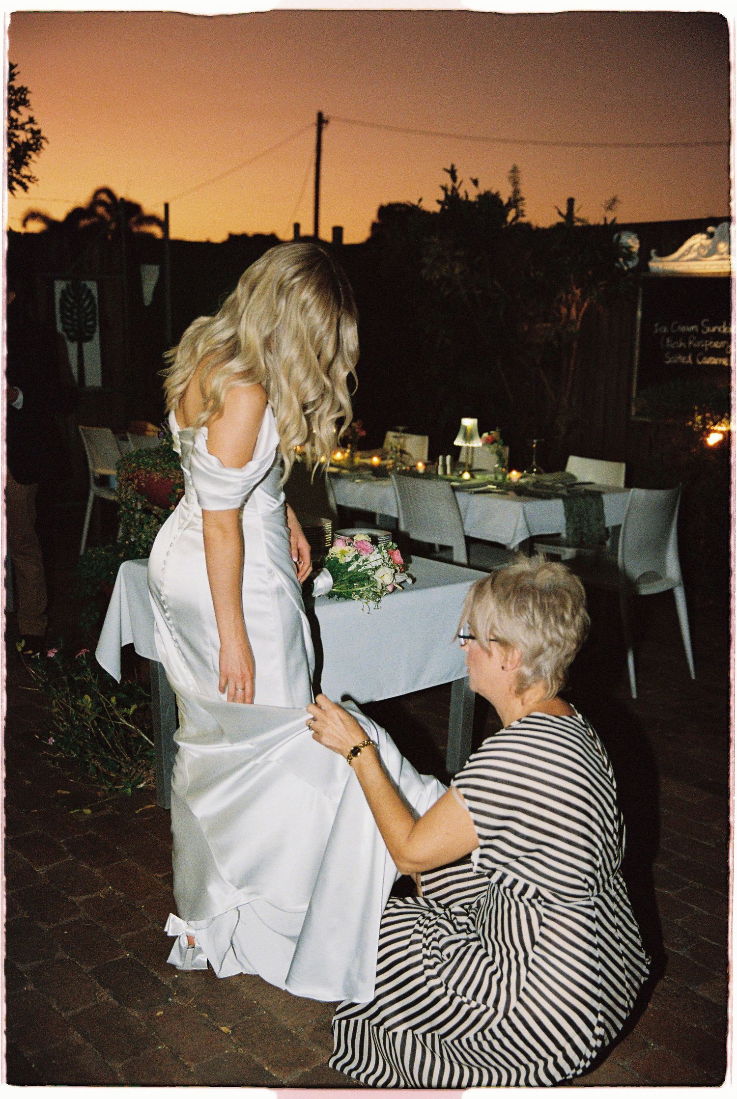Mother kneeling to adjust bride's white wedding gown train outdoors at sunset during golden hour.