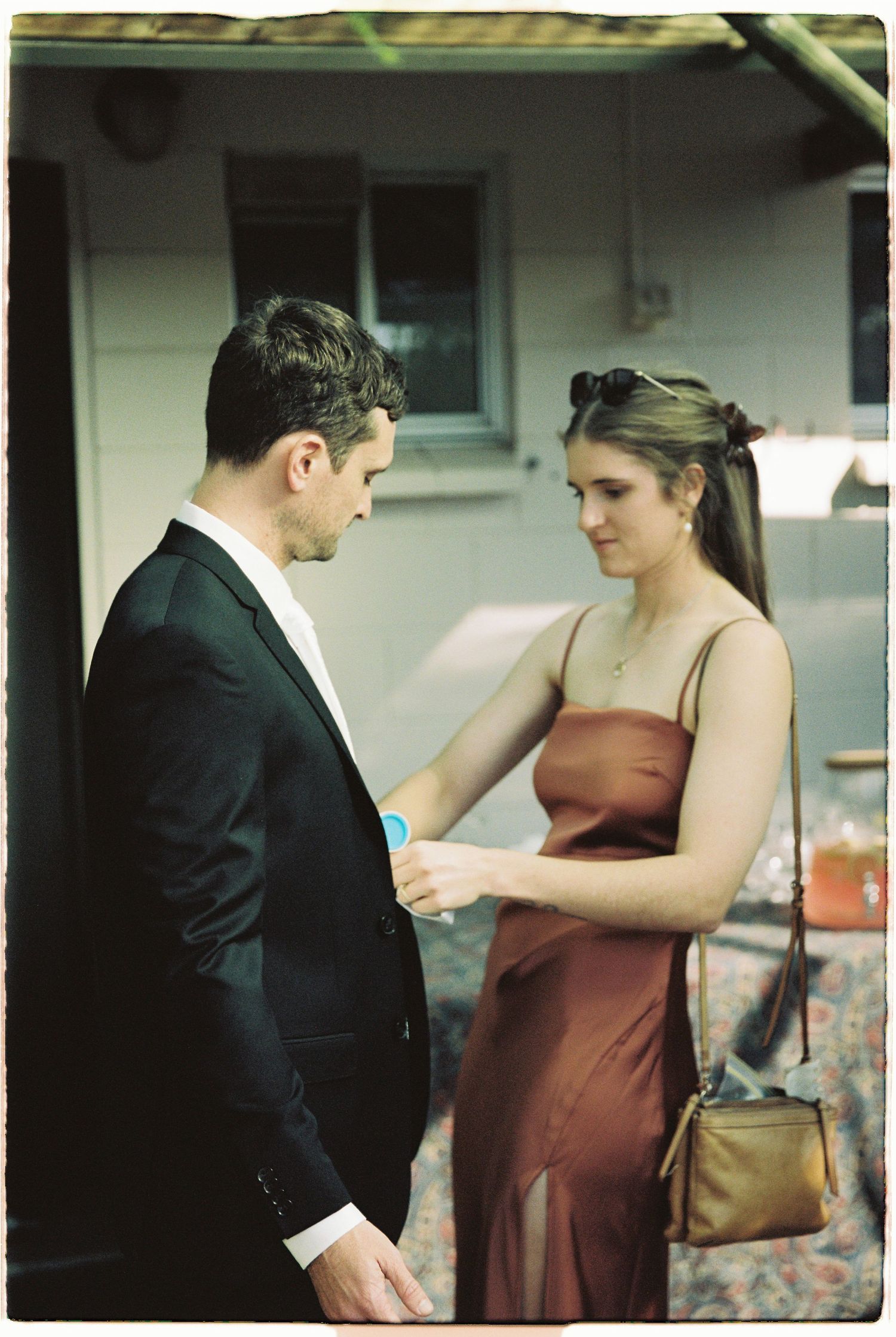 Man in black suit and woman in rust satin slip dress interact at outdoor wedding reception.