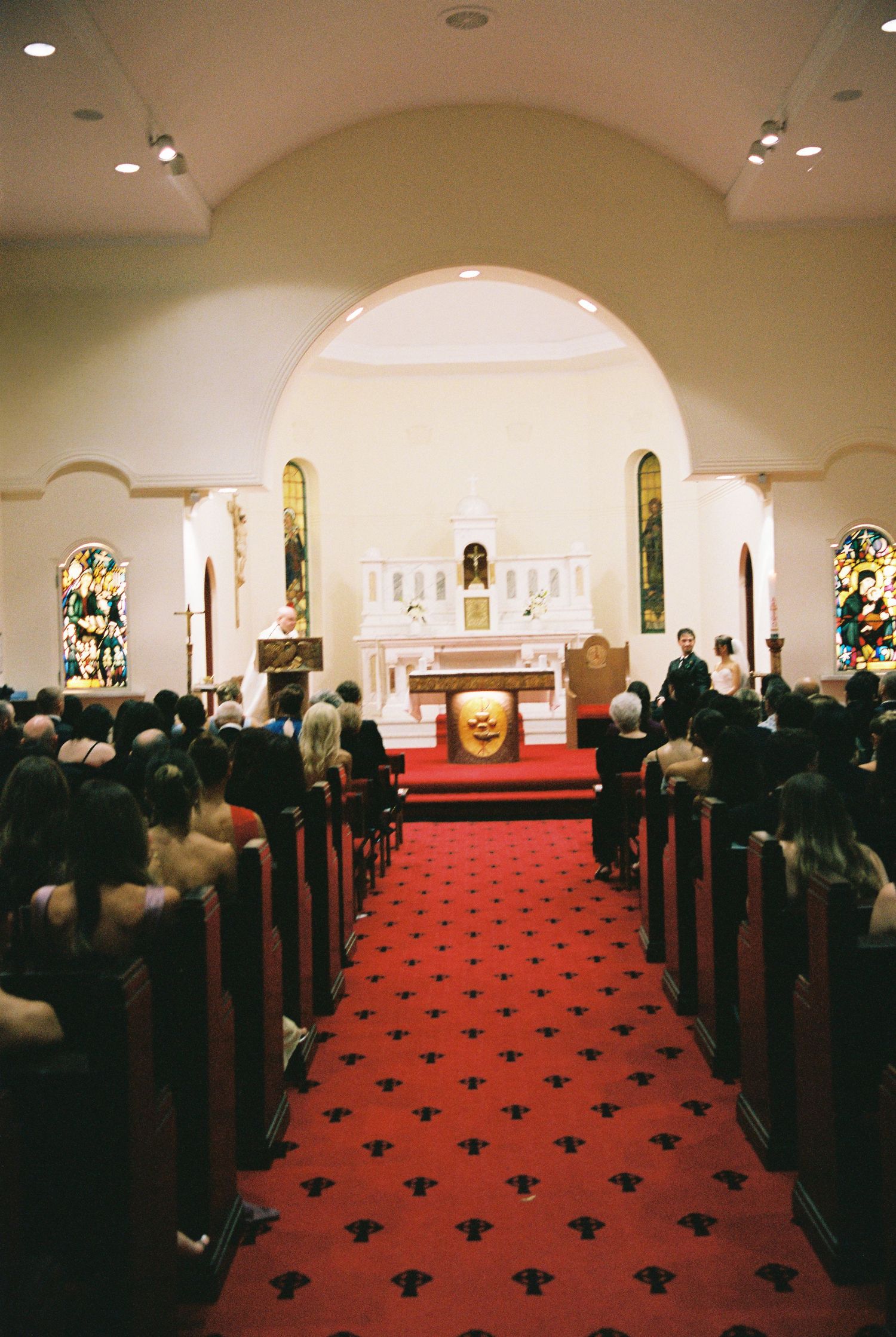 Wedding ceremony inside Catholic church with red carpet aisle, stained glass windows, and guests seated in wooden pews.