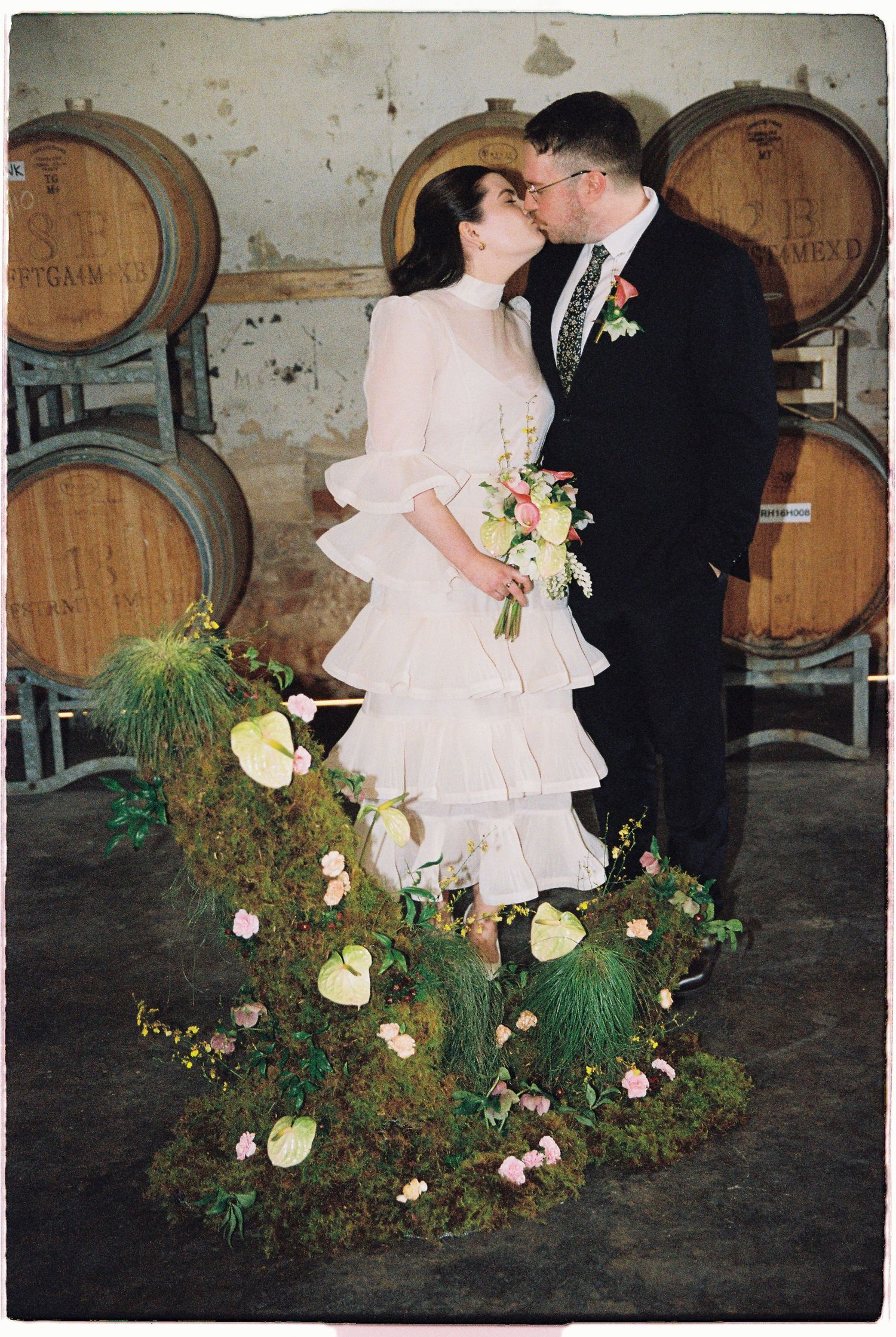 Bride and groom share a kiss at a winery wedding, surrounded by oak barrels and a lush floral moss installation.
