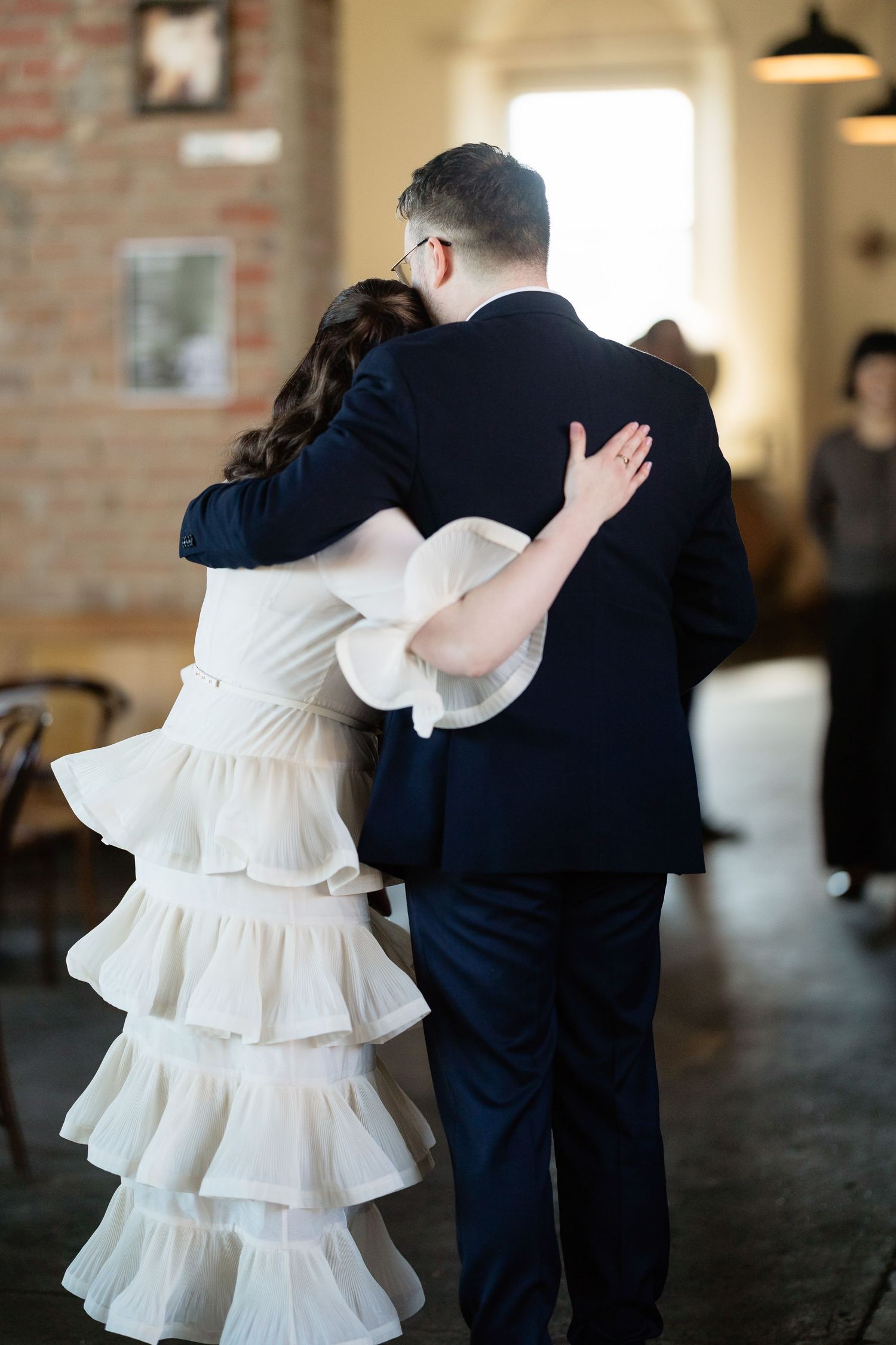 Bride in tiered white ruffled dress and groom in navy suit share first dance embrace at rustic wedding venue.