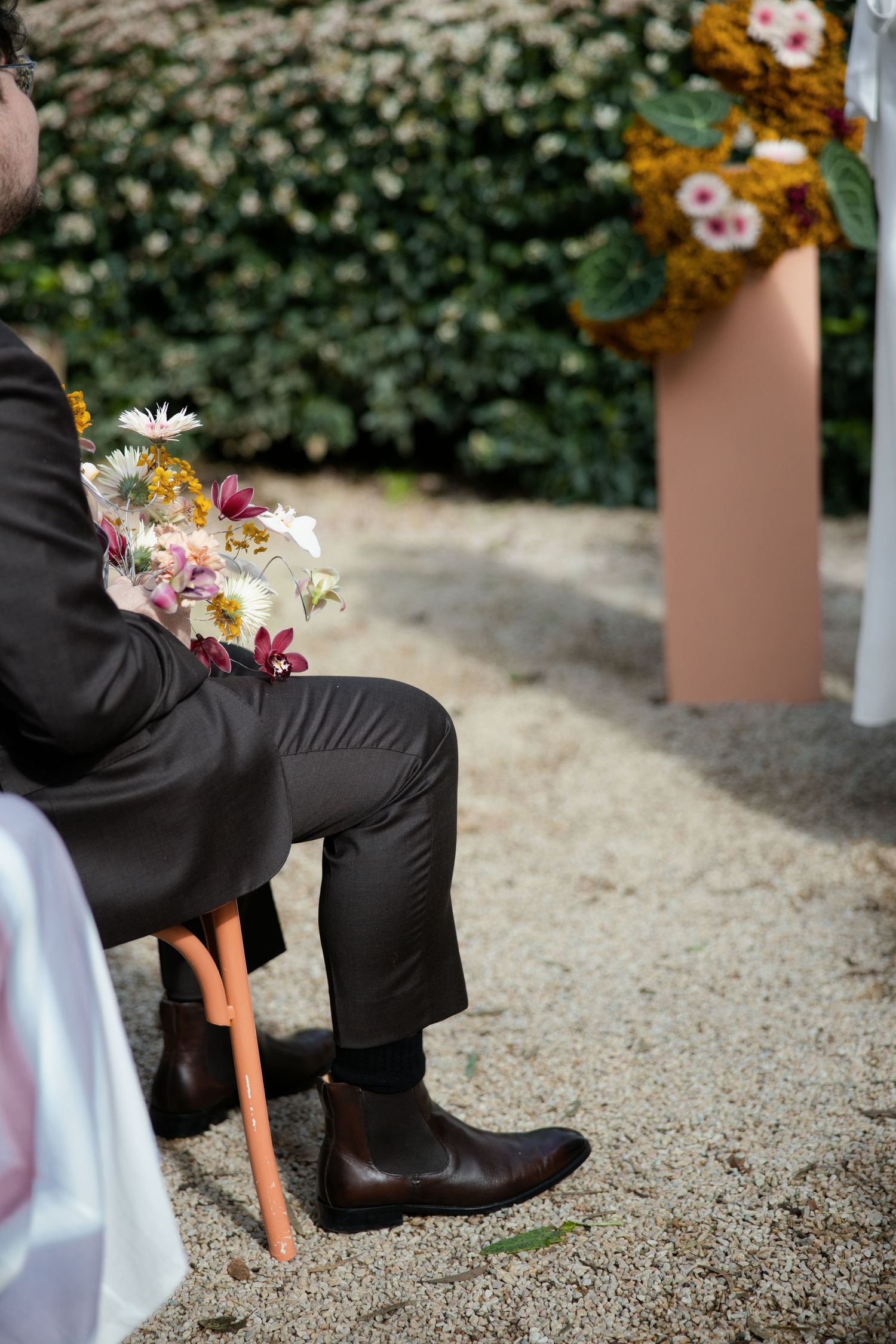 Man in dark suit holding wildflowers seated at outdoor wedding ceremony with floral arch and green hedges in background.