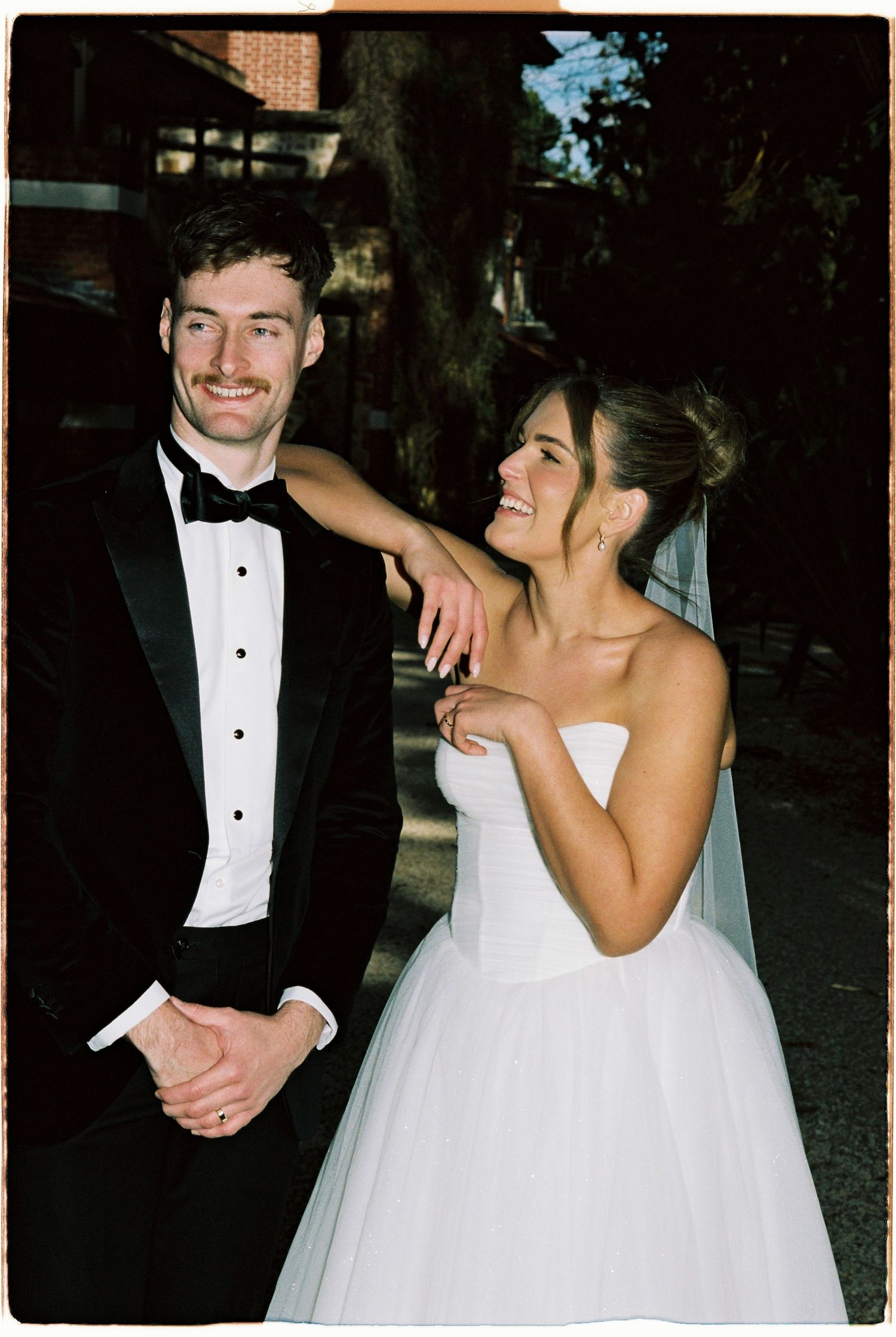 Smiling bride in strapless white ball gown and groom in black tuxedo posing together outdoors on their wedding day.