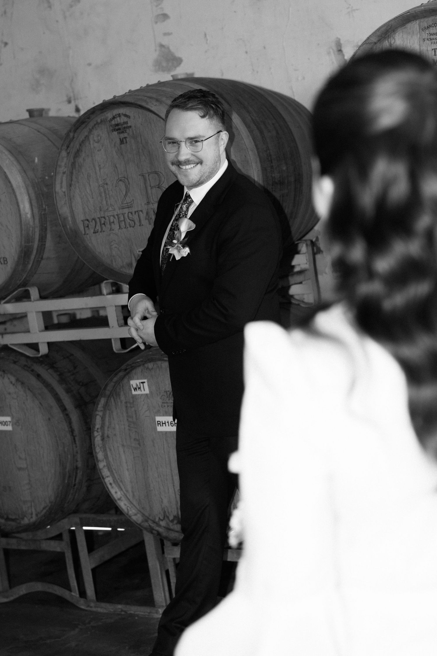 Groom in black suit smiles at bride near wine barrels in winery cellar, black and white wedding photo.