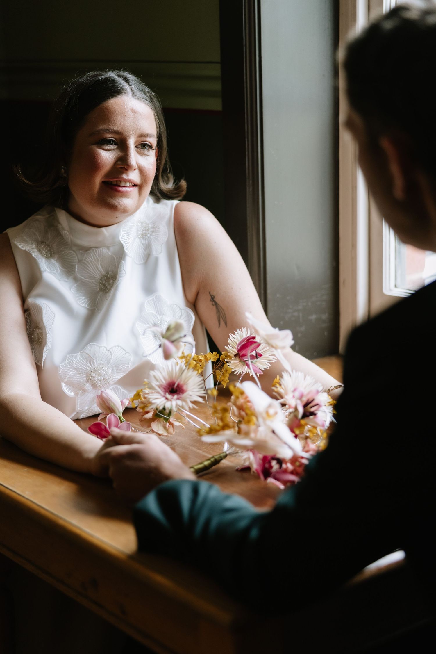 Smiling bride in white dress holds colorful floral bouquet while sitting at wooden table near window with groom.