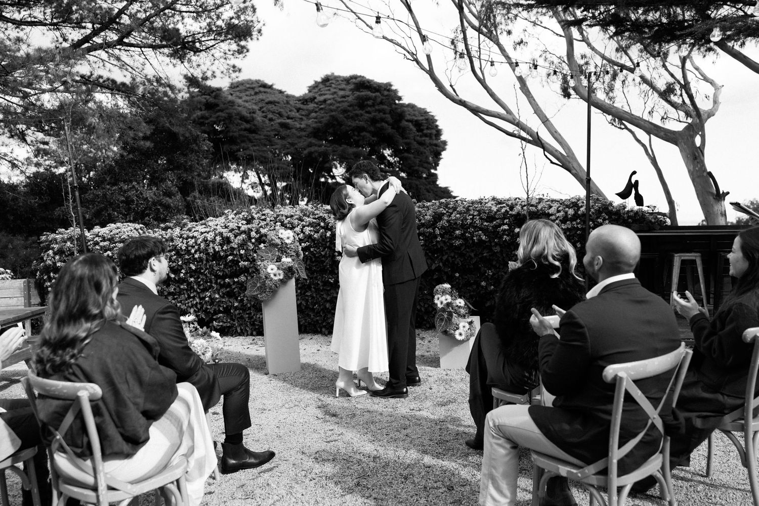 Black and white photo of a couple sharing their first kiss at an outdoor wedding ceremony with seated guests applauding.