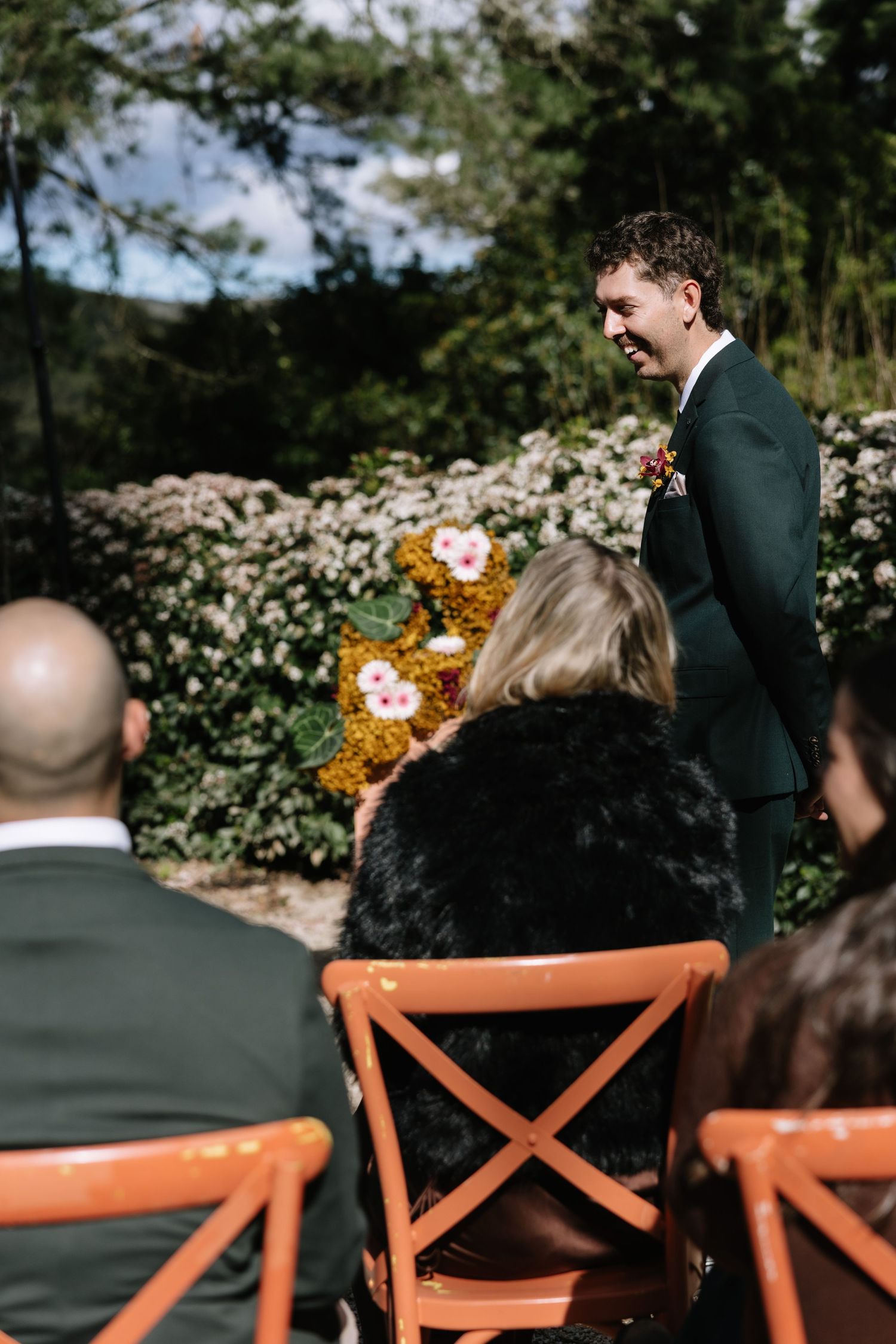 Groom in dark green suit smiling at wedding guests seated in orange chairs outdoors, surrounded by lush floral arrangements.