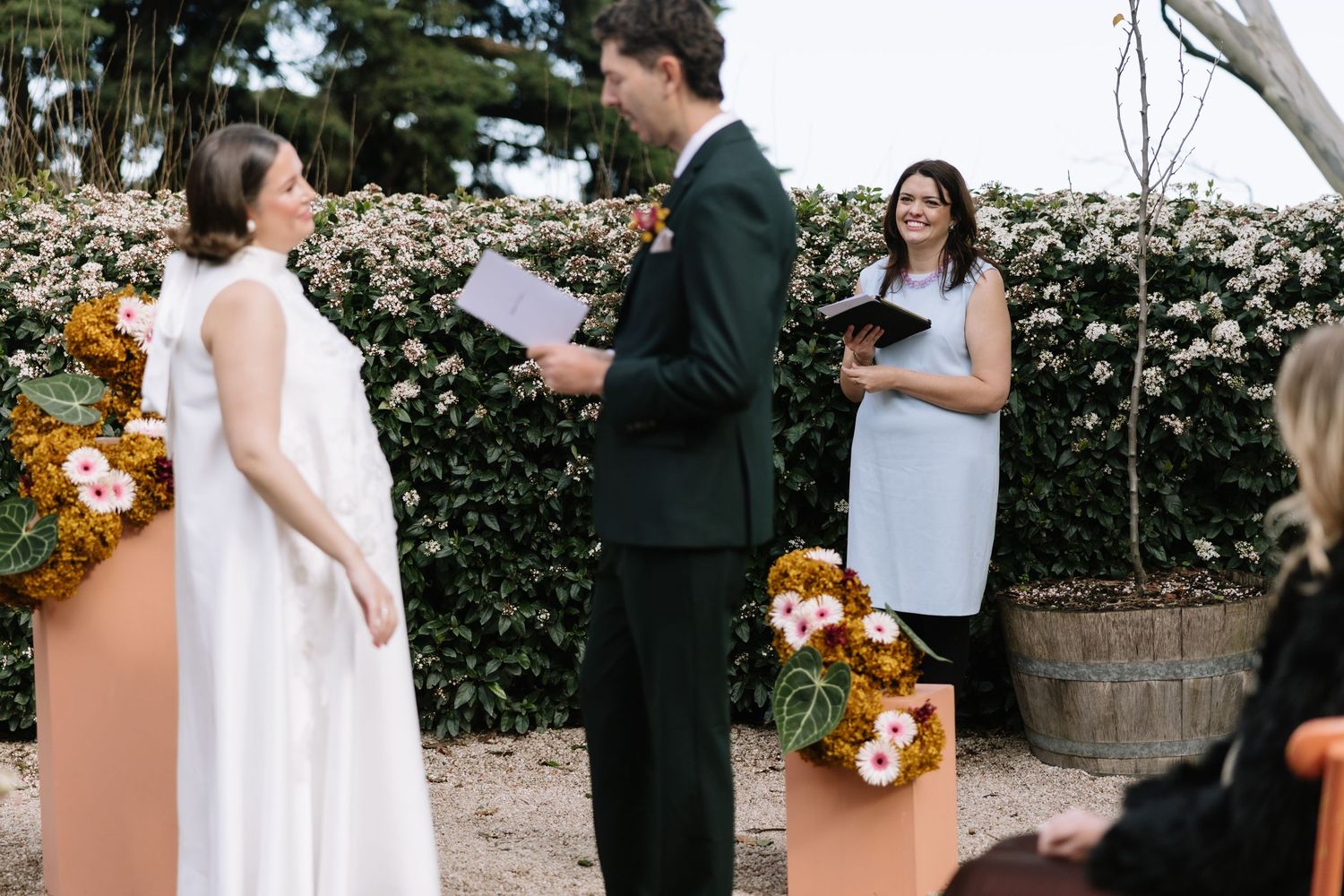 Outdoor wedding ceremony with bride in white dress, groom in dark suit, and officiant holding papers, surrounded by floral arrangements.