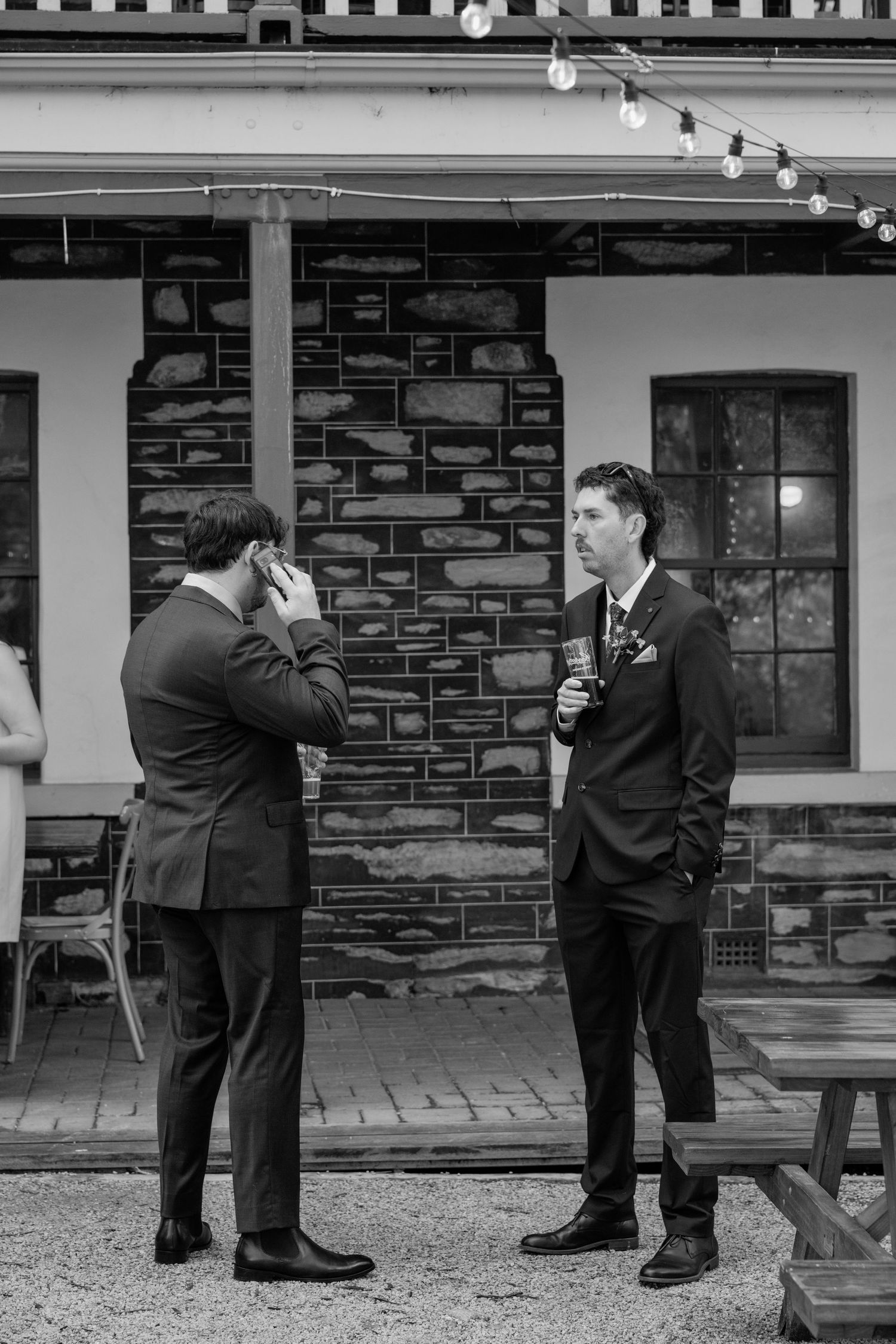 Two grooms in suits share a moment outside a stone building at their wedding reception.