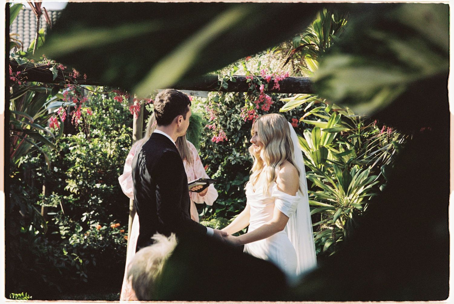 Bride and groom exchange vows in an intimate outdoor garden ceremony framed by tropical foliage.
